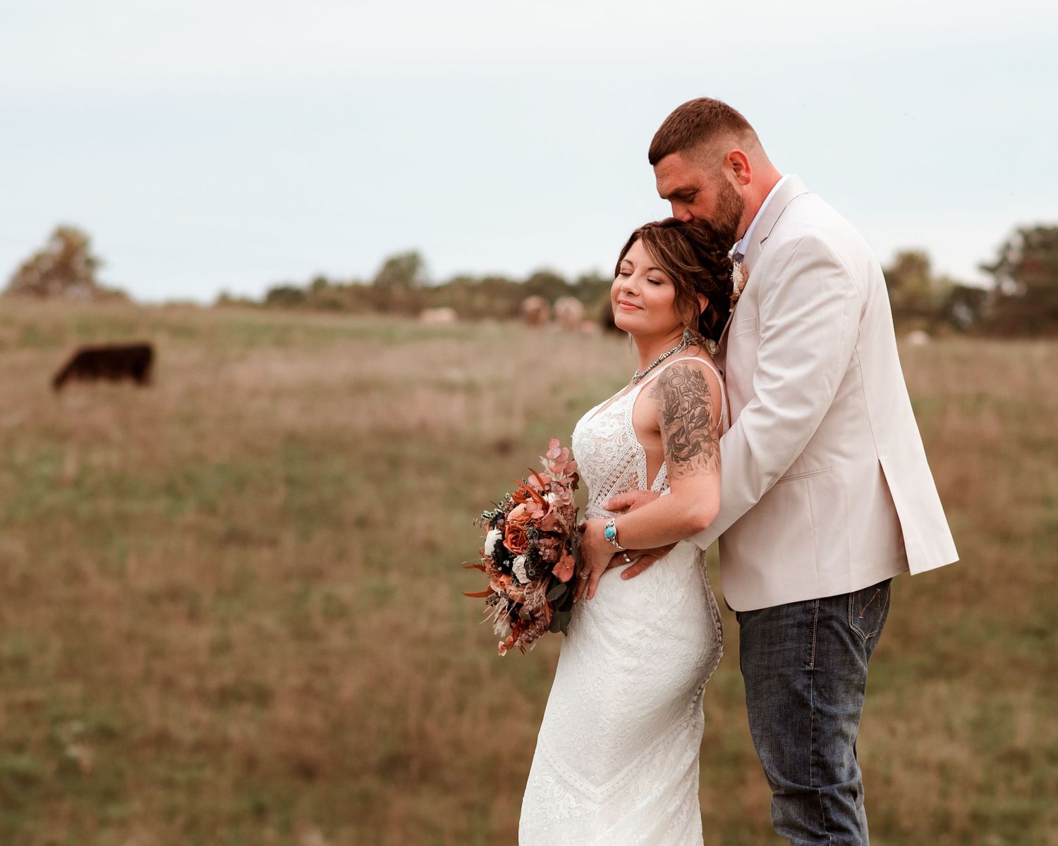 Bride and groom embrace in a rustic field, bride holding an autumn-toned bouquet with a cow grazing in the background.