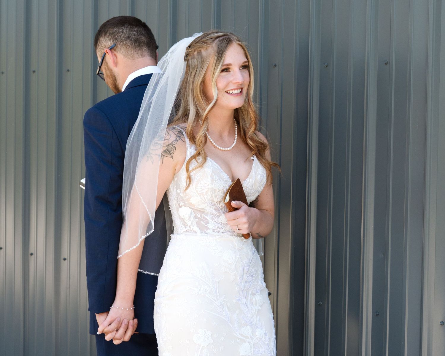 Smiling bride in lace wedding gown and veil holds groom's hand outdoors near metal wall.