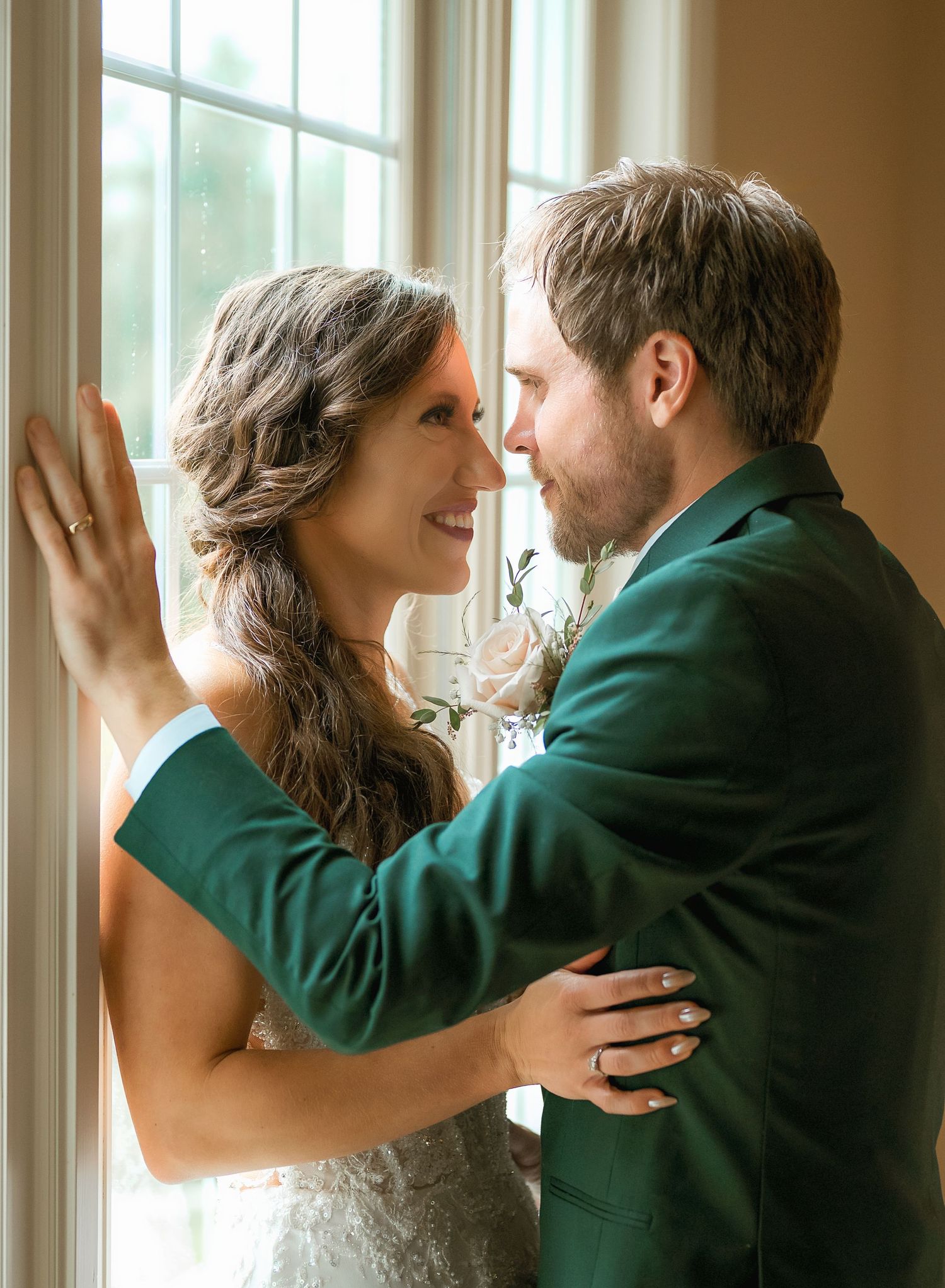 Bride and groom share an intimate moment by a sunlit window, groom in a dark green suit smiling.