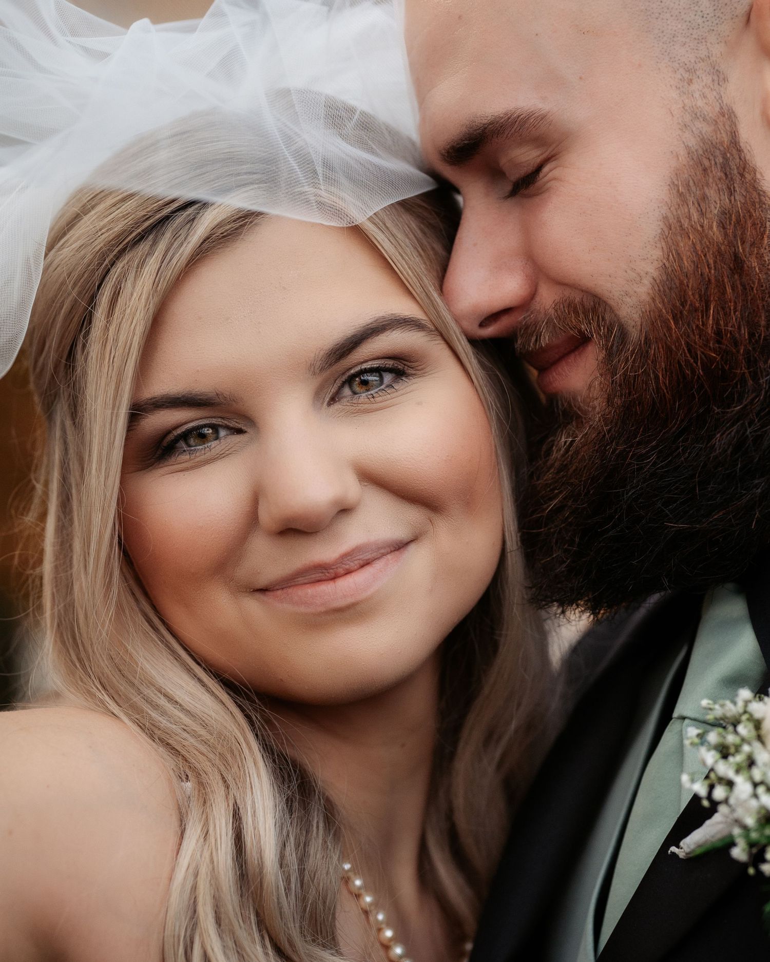 Close-up of smiling bride with green eyes and pearl necklace with bearded groom nuzzling her cheek.