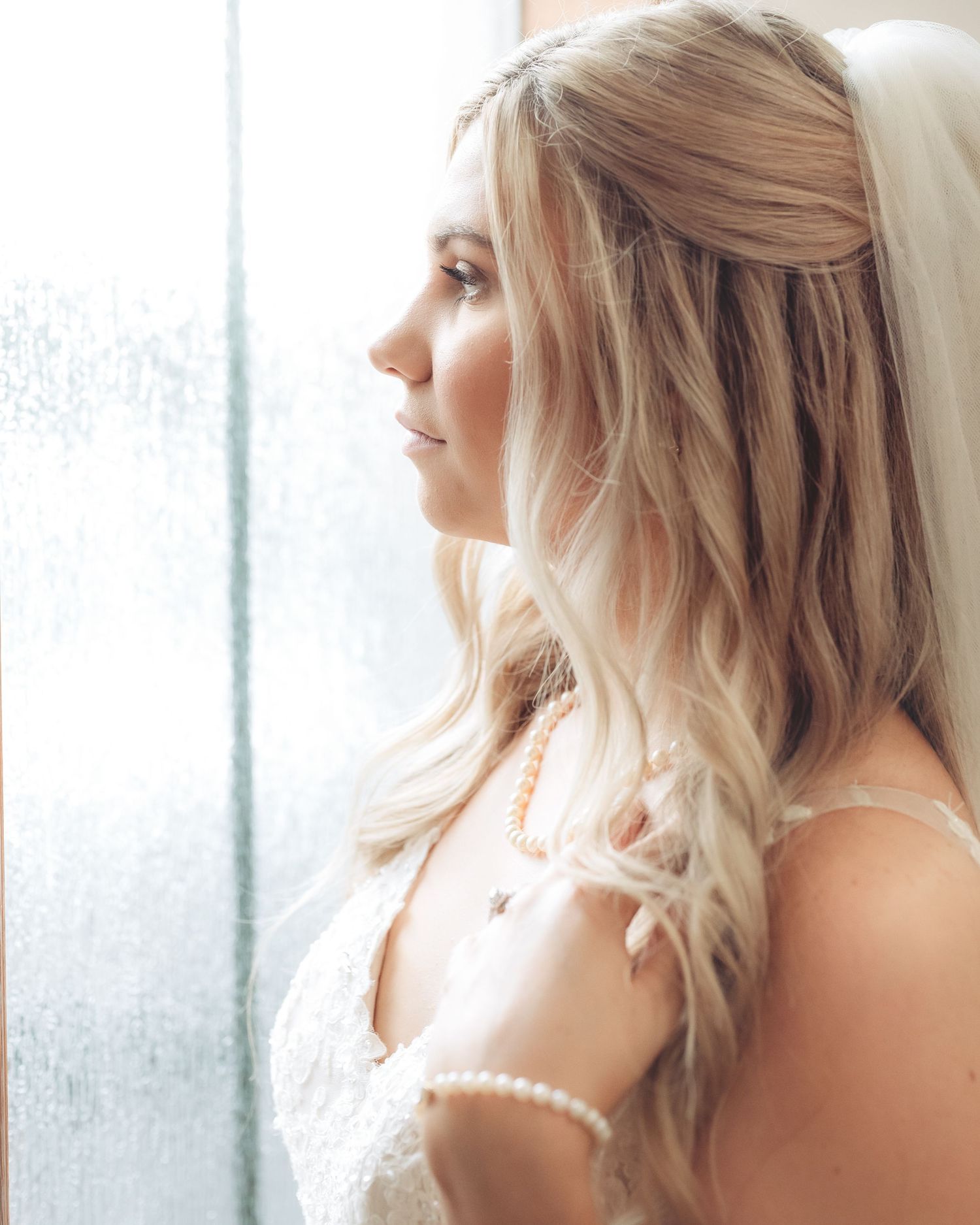 Elegant bride with blonde hair in lace gown and pearl necklace gazes out a window on her wedding day.