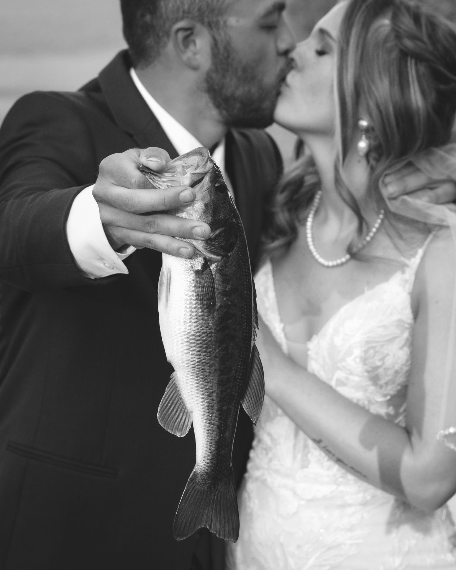 Black and white photo of bride and groom kissing while holding a large bass fish on their wedding day.