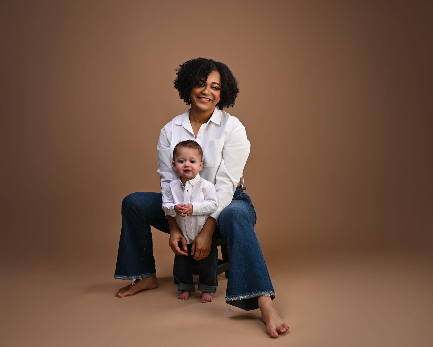 A mother in white shirt sits smiling with her toddler boy on a warm tan studio background.