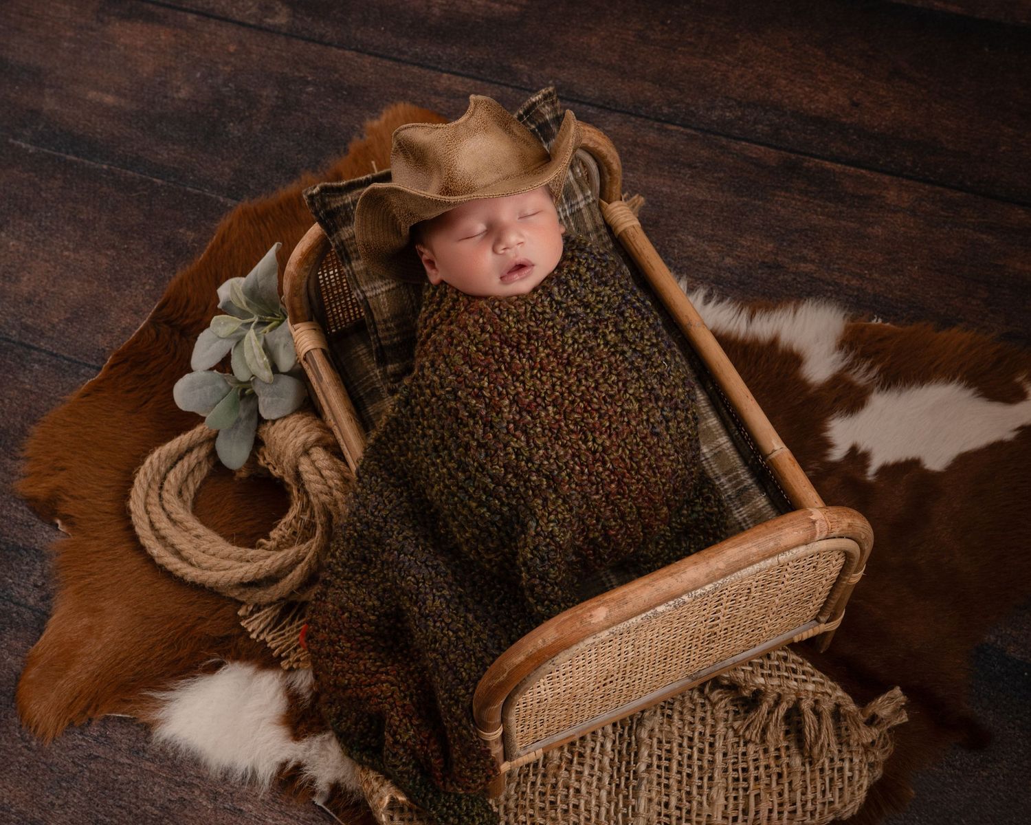 Sleeping newborn wrapped in brown knit wearing a cowboy hat on a cowhide rug in a wooden crib.