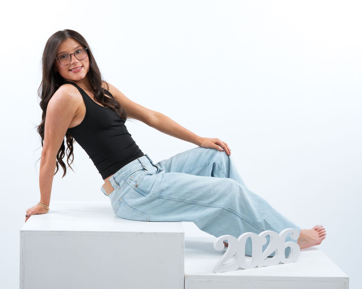 Young woman in black tank top and jeans sitting on white steps next to 2026 sign.