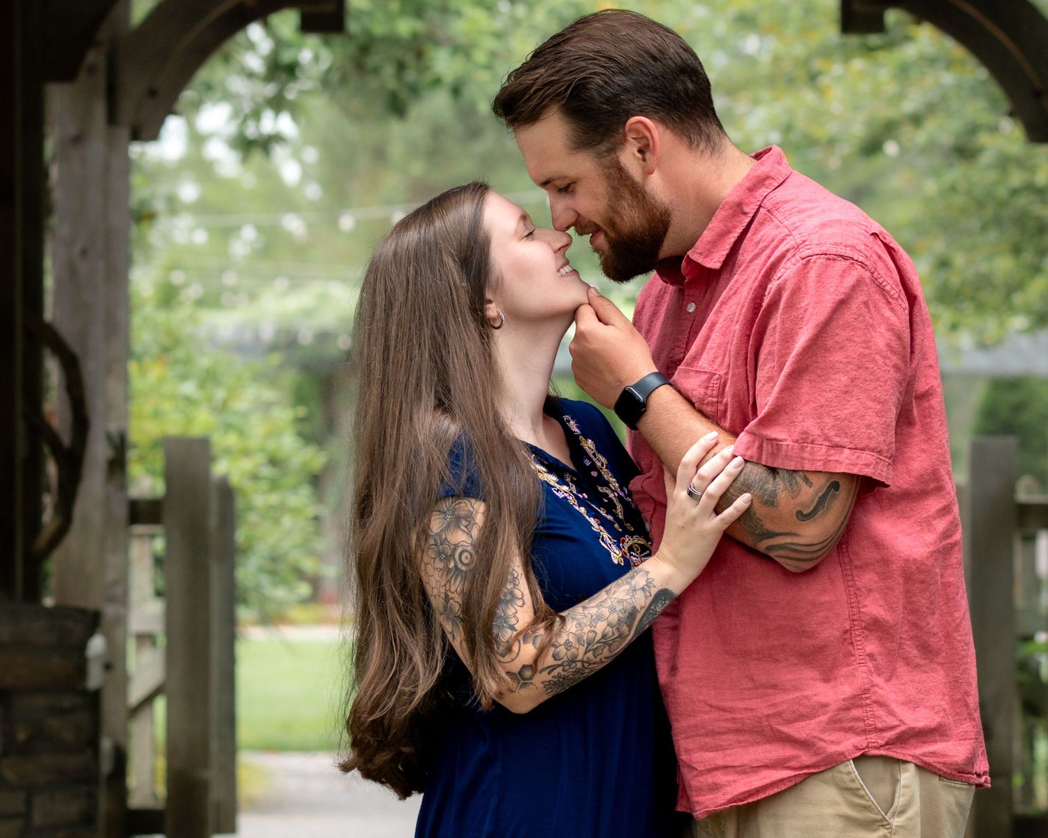 Tattooed couple sharing a romantic moment under a wooden pergola in a lush garden setting.
