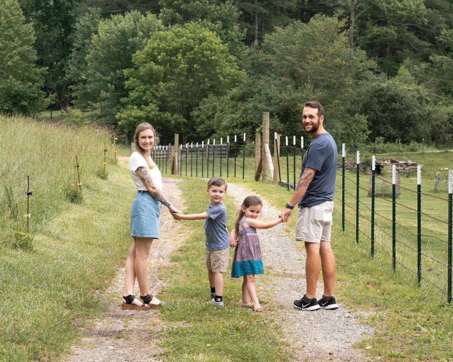 A joyful family of four holds hands along a gravel path surrounded by green countryside scenery.