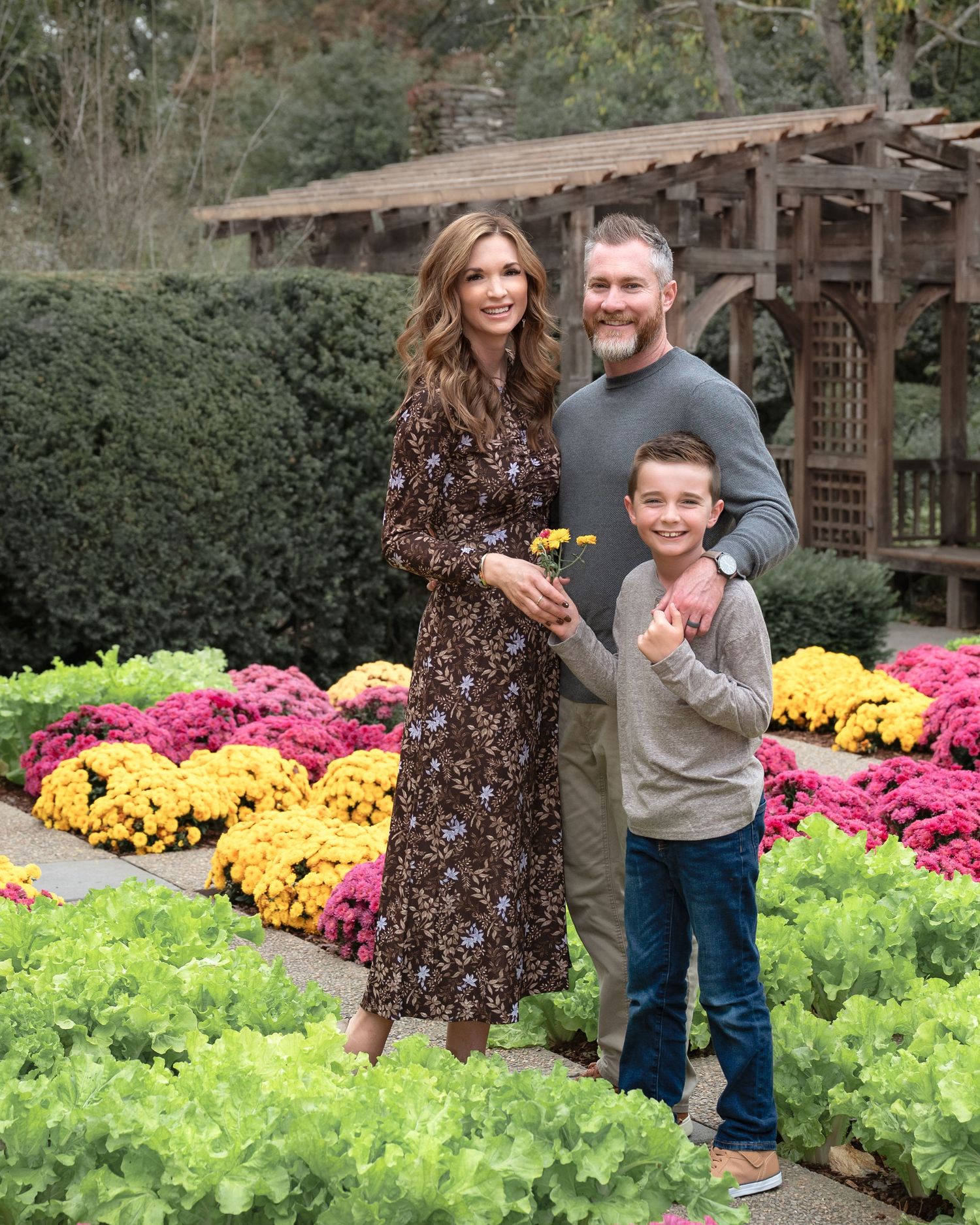 A family of three poses together in a garden path surrounded by colorful chrysanthemums.