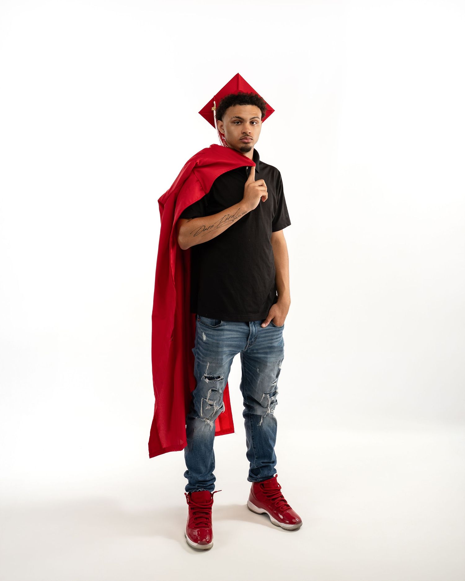 Male graduate in red cap and gown stands confidently in studio wearing red Jordan sneakers.
