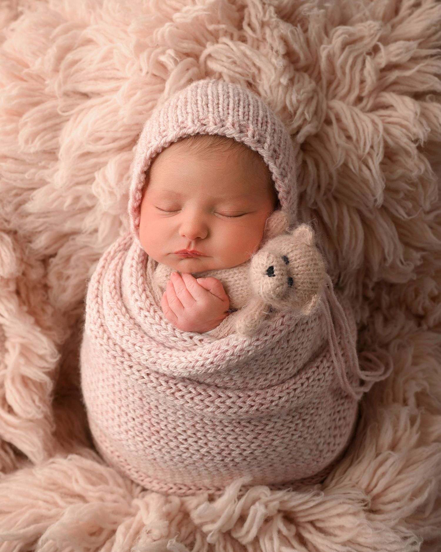 Newborn baby in pink knit wrap and bonnet sleeping with a small teddy bear on fluffy rug.