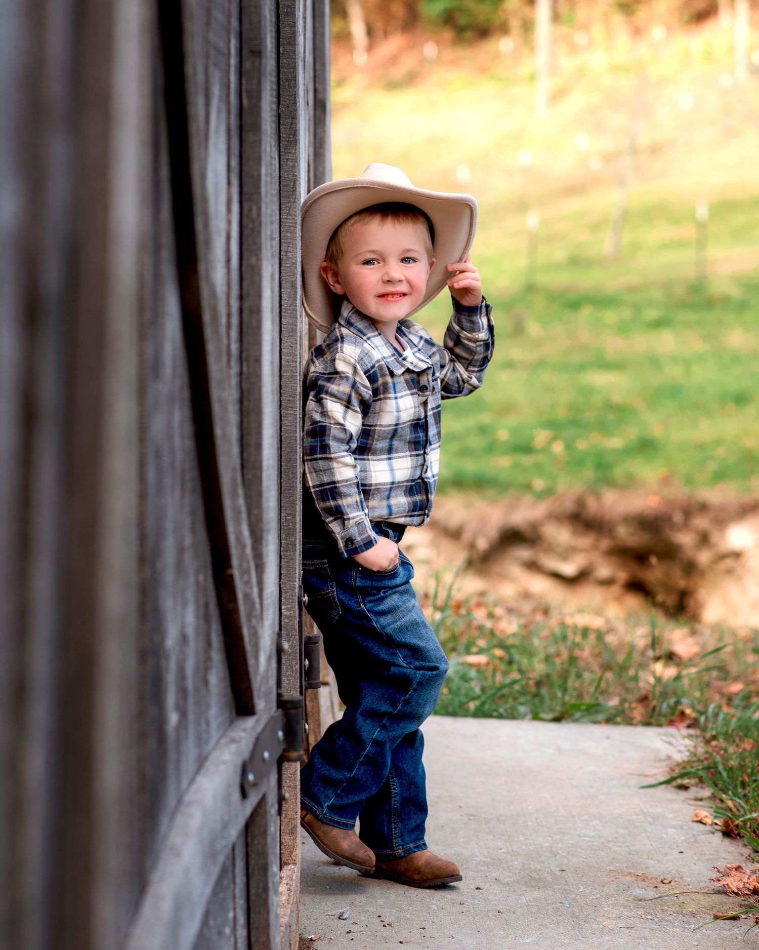Adorable toddler boy in cowboy hat and boots leaning against a barn door smiling.