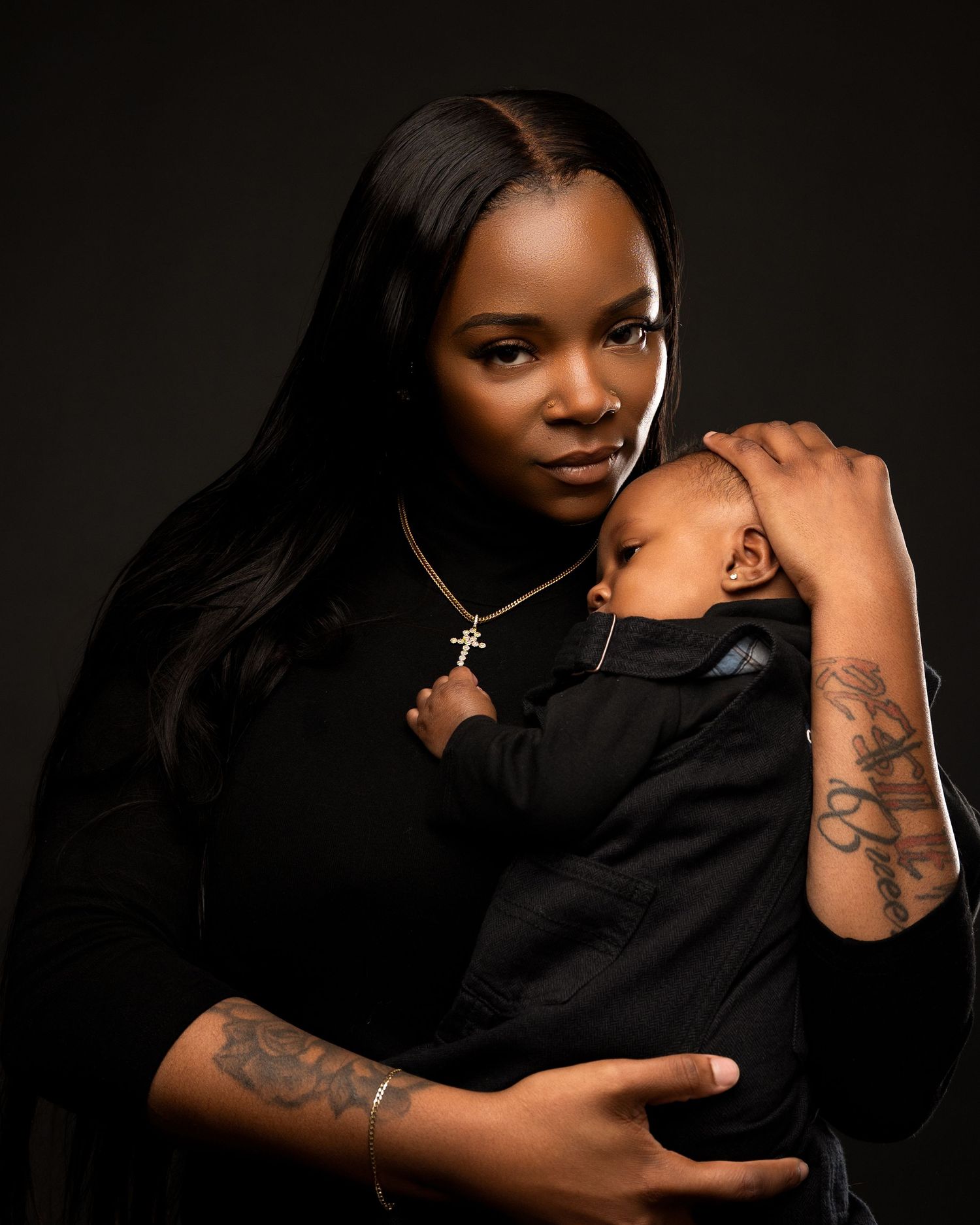 A mother in black holds her sleeping newborn close in a dramatic dark studio portrait.