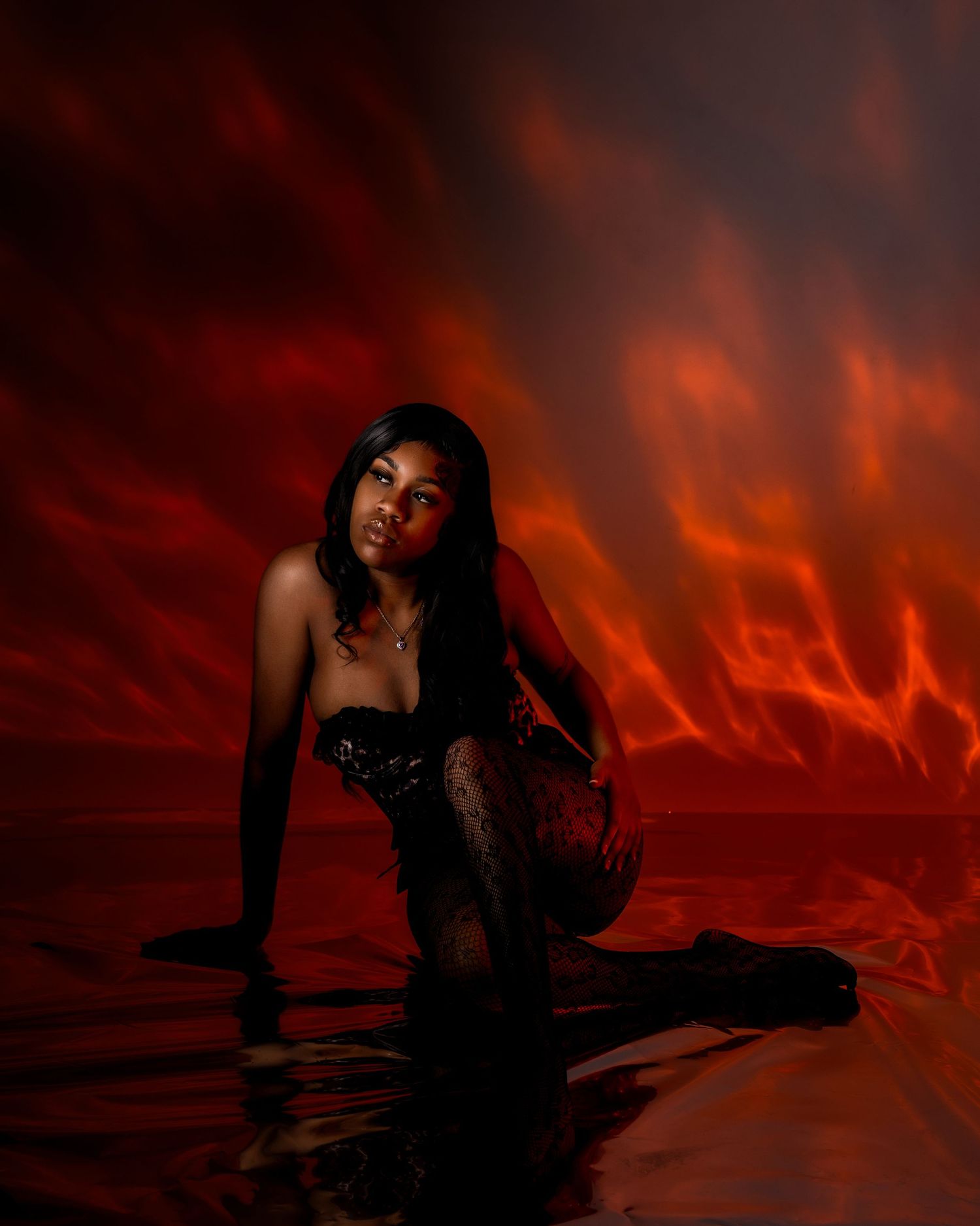 Woman in black lace lingerie and heeled boots kneeling on a glossy reflective floor against a dramatic red flame-patterned backdrop.