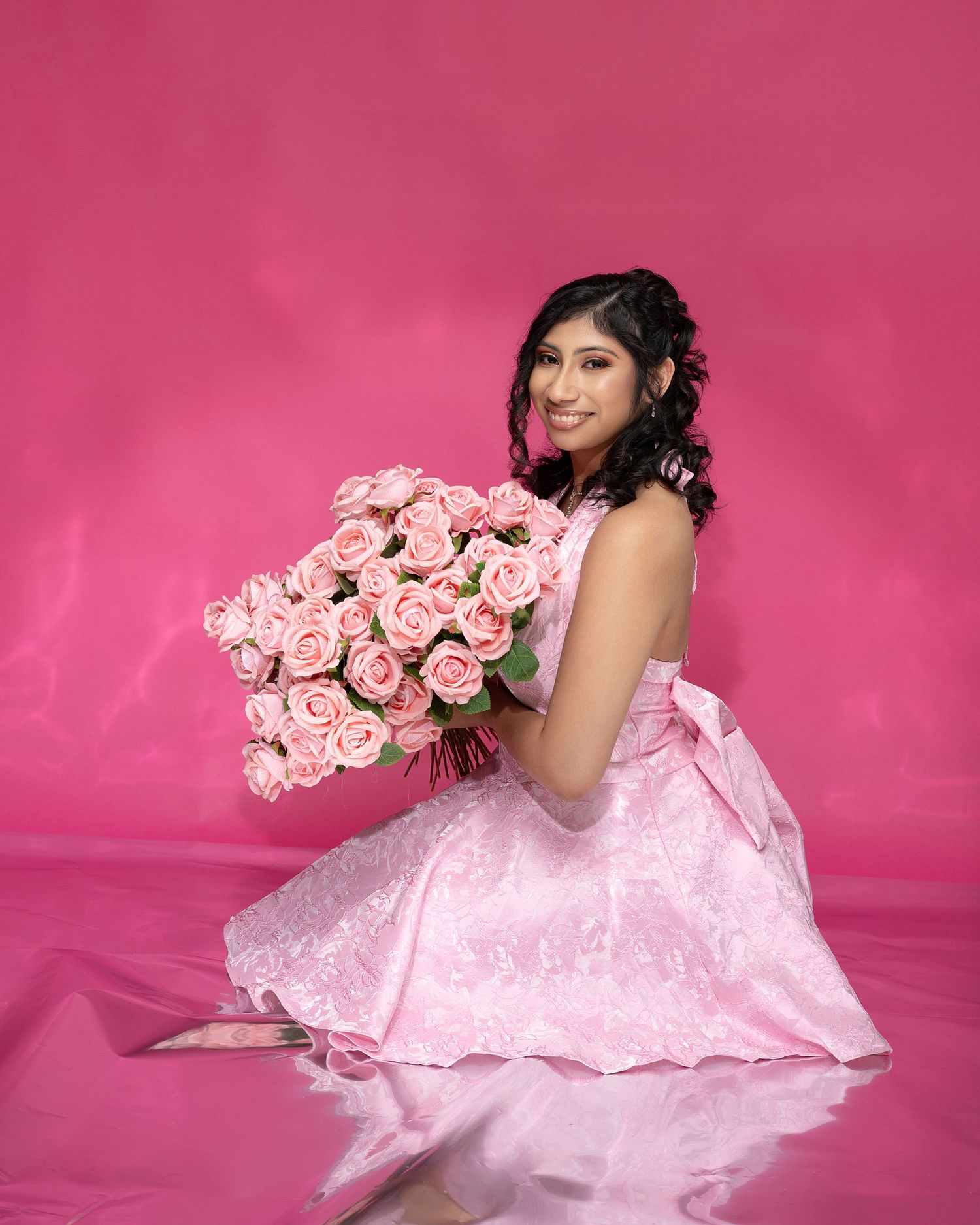 Woman in a pink lace dress kneeling on a glossy pink floor, smiling while holding a large bouquet of pink roses against a pink backdrop.