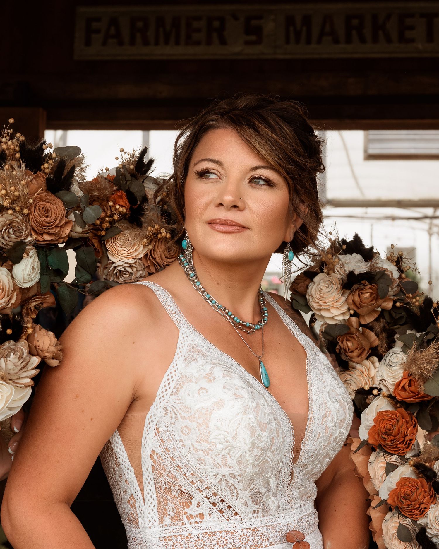 Bride in lace wedding gown posing at Farmer's Market with rustic floral arrangements in warm earth tones.