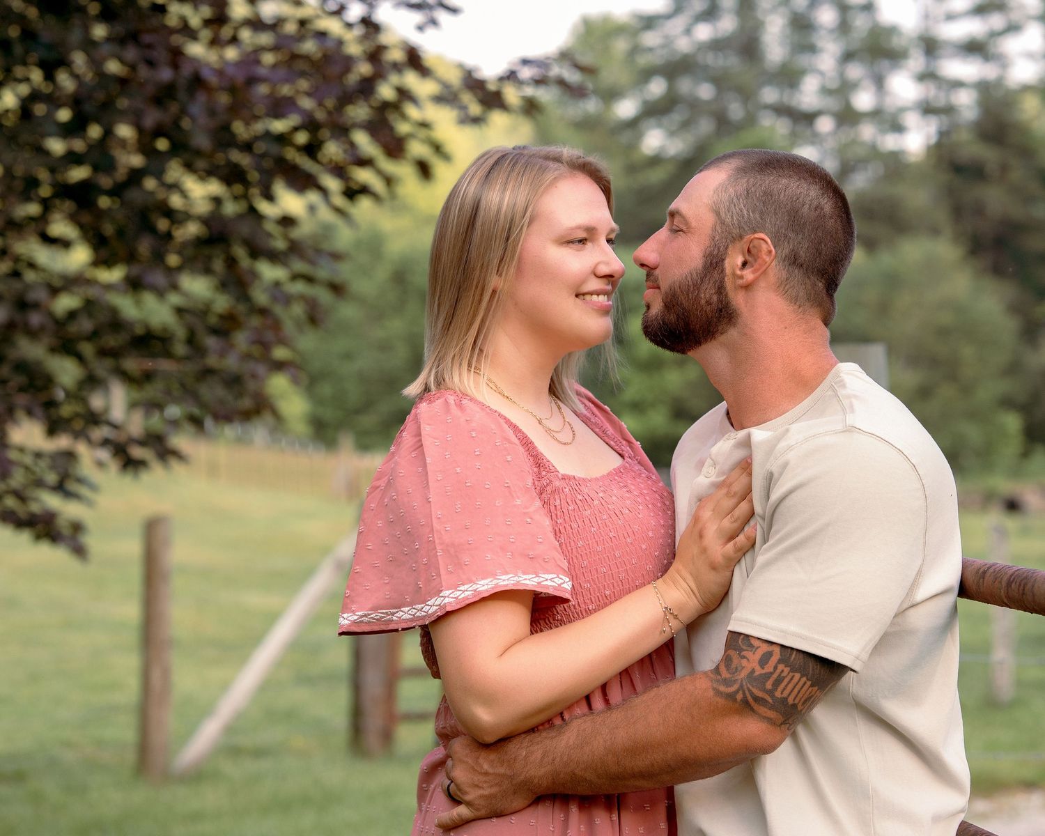 A young couple shares a romantic moment outdoors near a wooden fence in green fields.