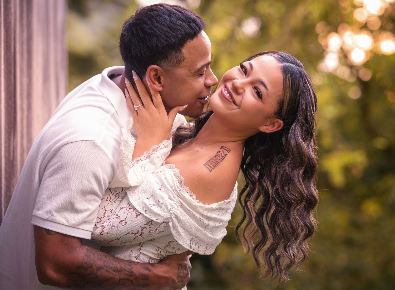 Intimate couple moment outdoors, woman in white lace dress smiling as man kisses her cheek near greenery.