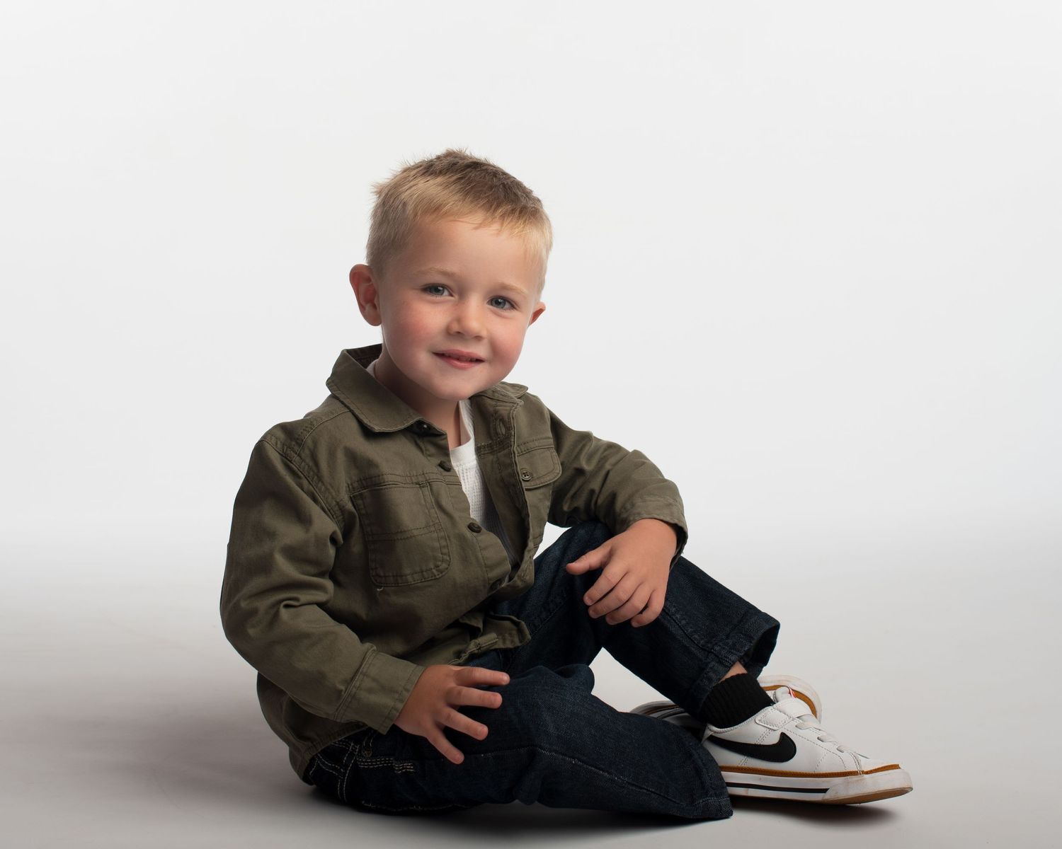 Cheerful young blonde boy in olive shirt and jeans sitting casually on white studio floor smiling.