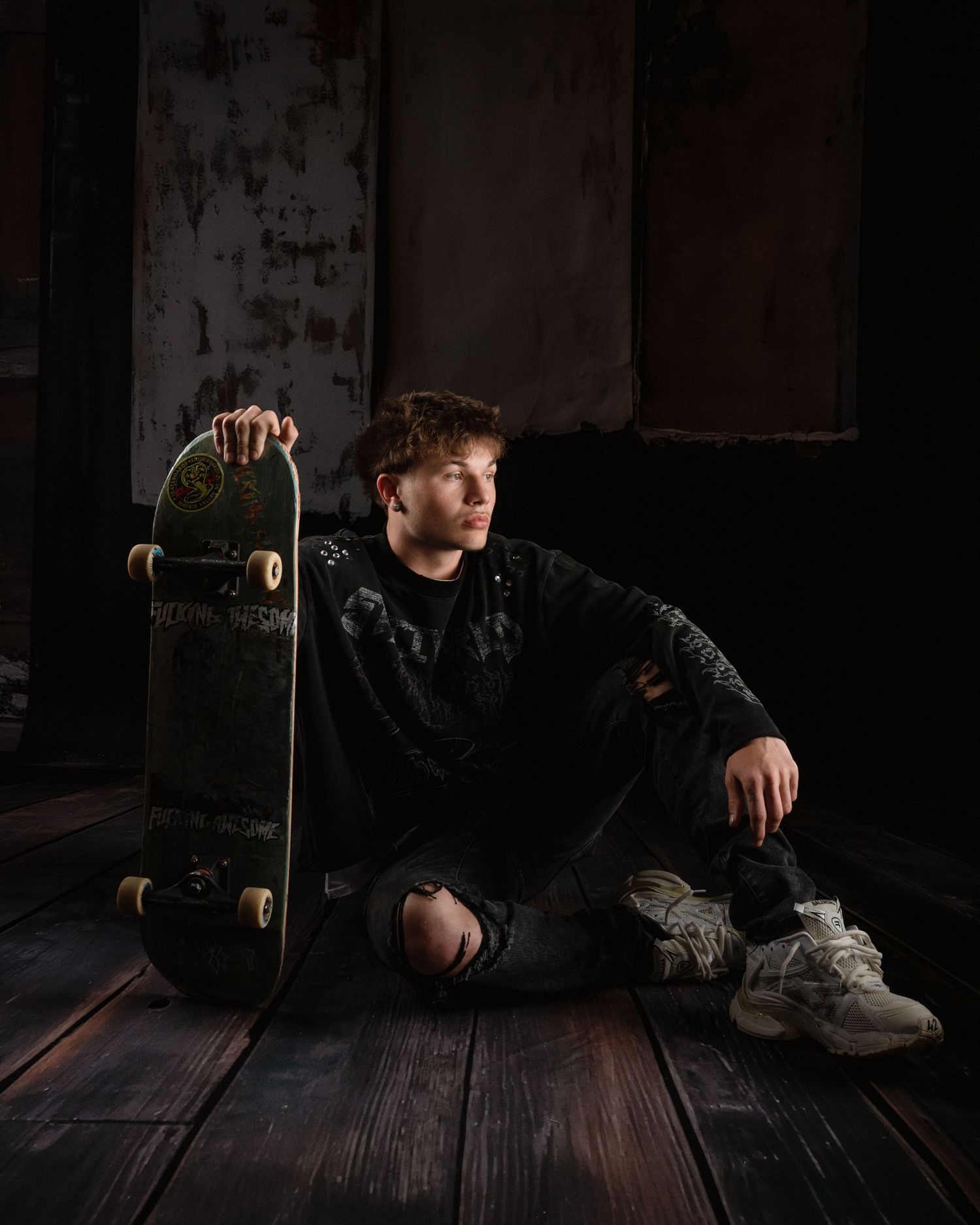 Young man in black ripped jeans sitting on wooden floor holding skateboard in dark urban setting.
