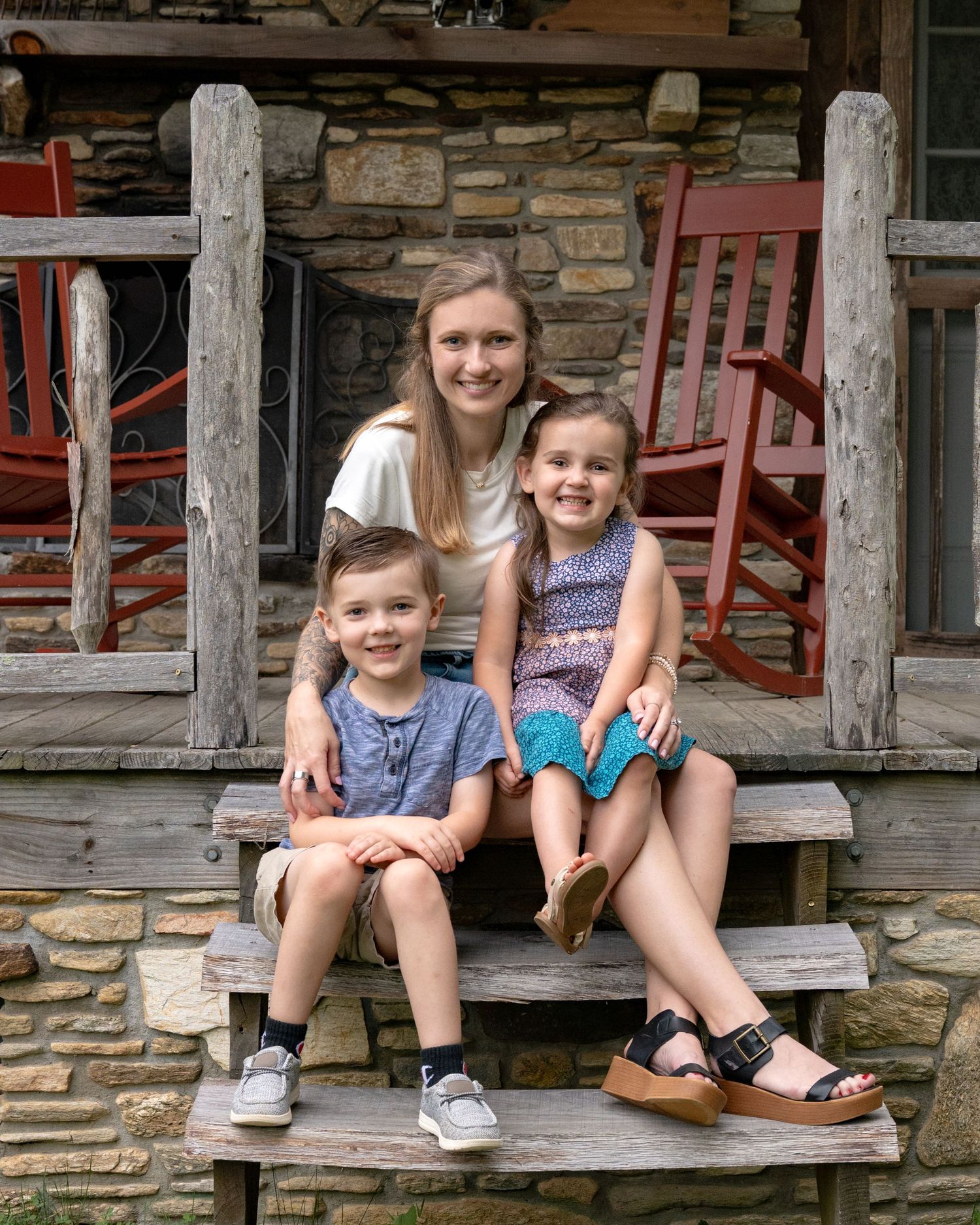 A teenage girl poses with two young children on rustic stone cabin steps with red rocking chairs.