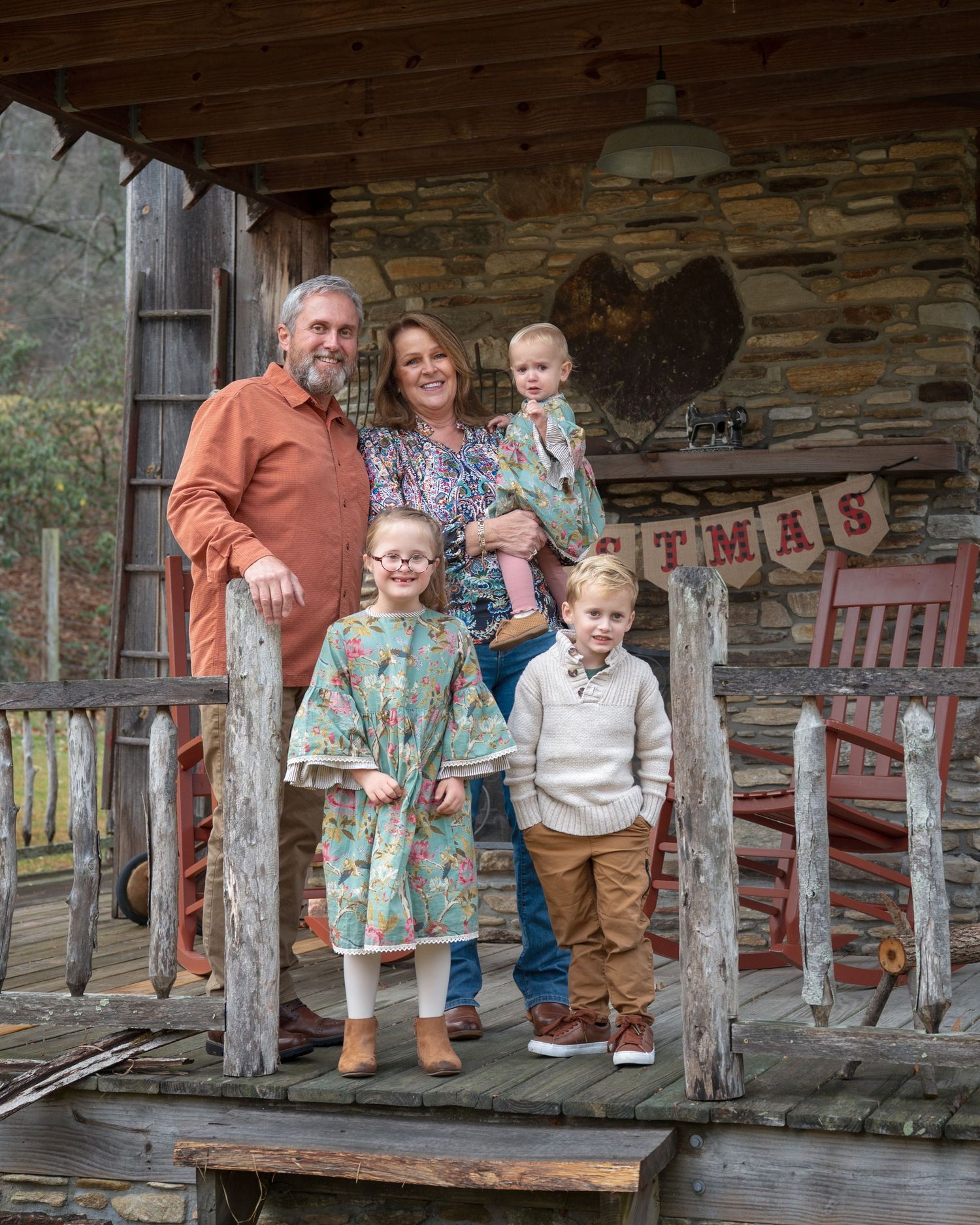 A multigenerational family poses on a stone cabin porch with a Christmas banner backdrop.
