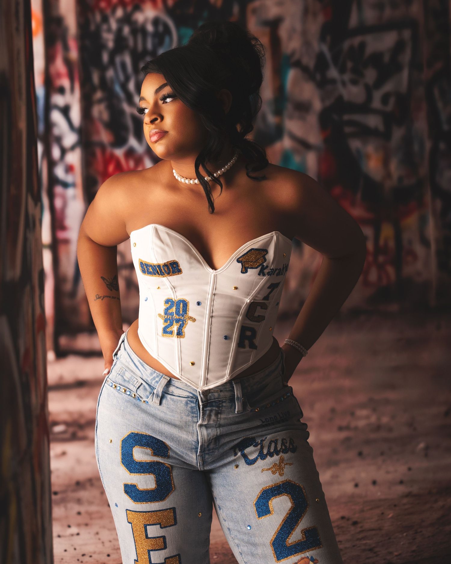 Senior girl poses in custom Class of 2027 corset and jeans in front of graffiti wall.