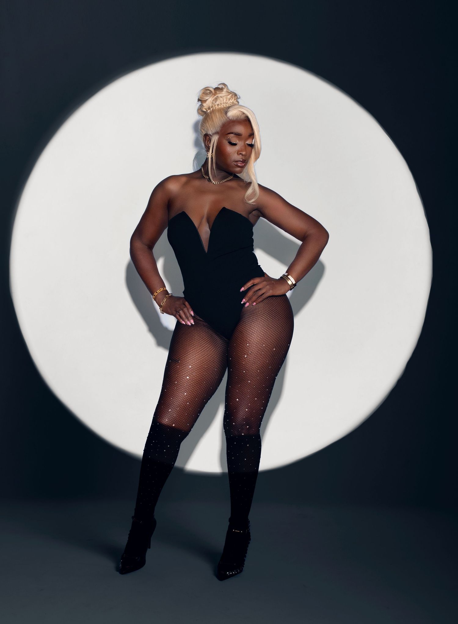 Woman in black bodysuit and fishnet tights posing confidently in front of circular spotlight backdrop.