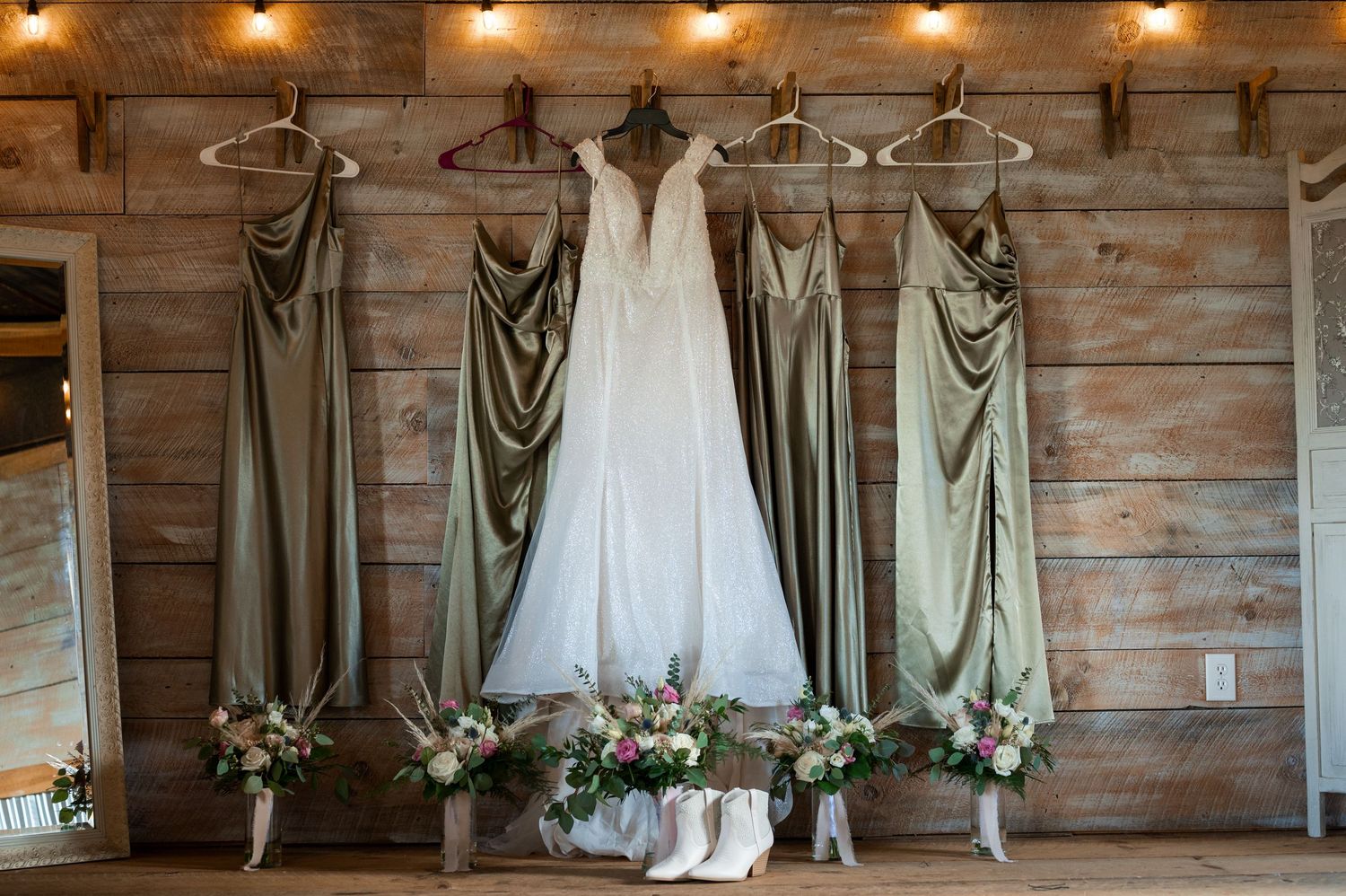 Wedding dress and sage green bridesmaid gowns hanging in rustic barn with floral arrangements.