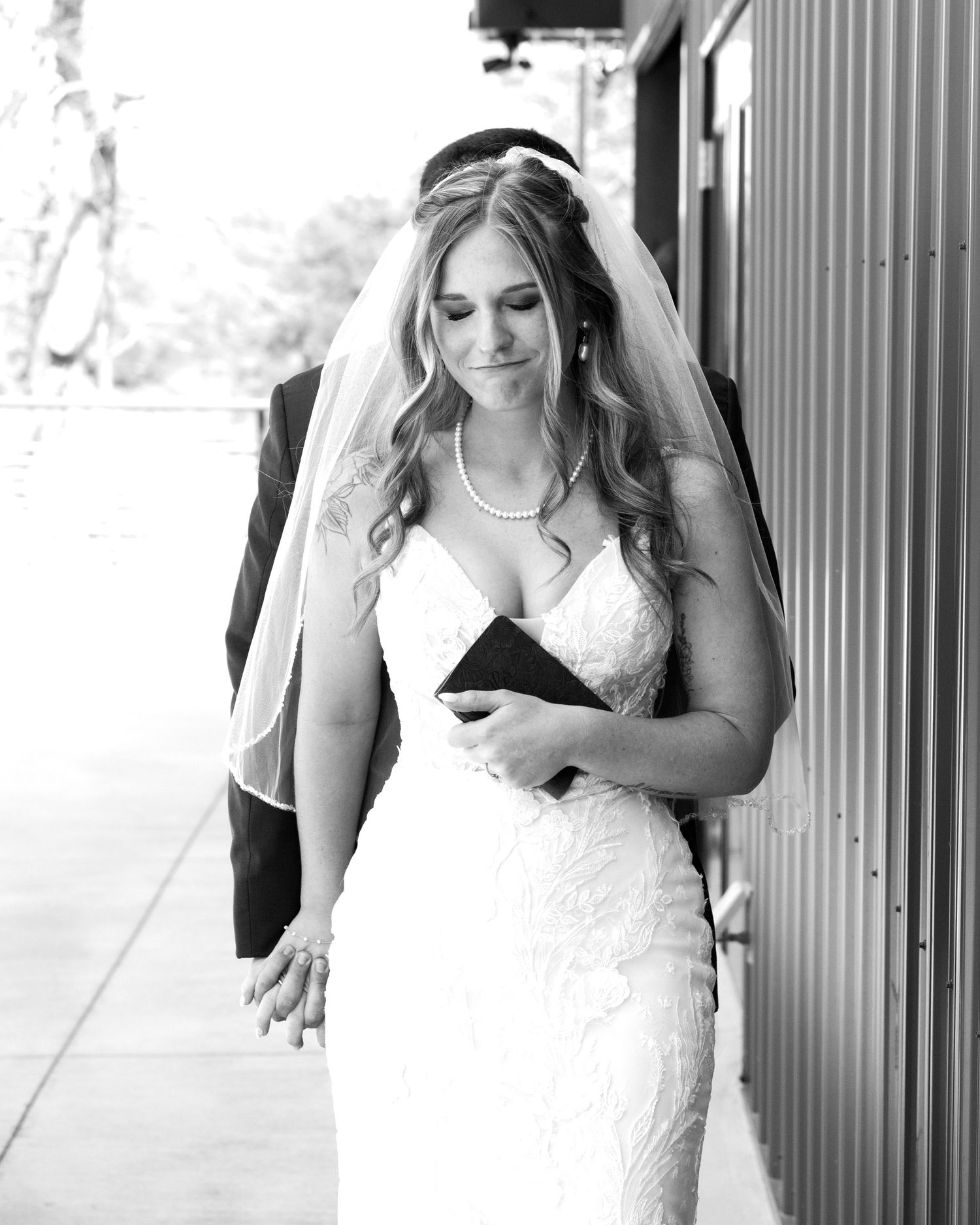 Black and white photo of bride in lace gown and veil walking with groom while holding a clutch purse.