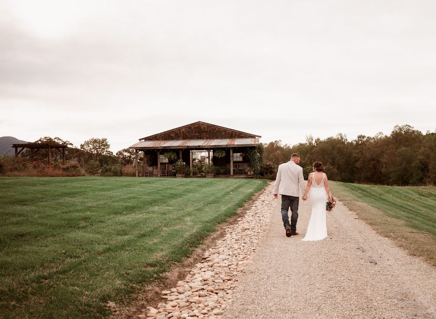 Bride and groom walking hand in hand down gravel path toward rustic barn venue on green estate.