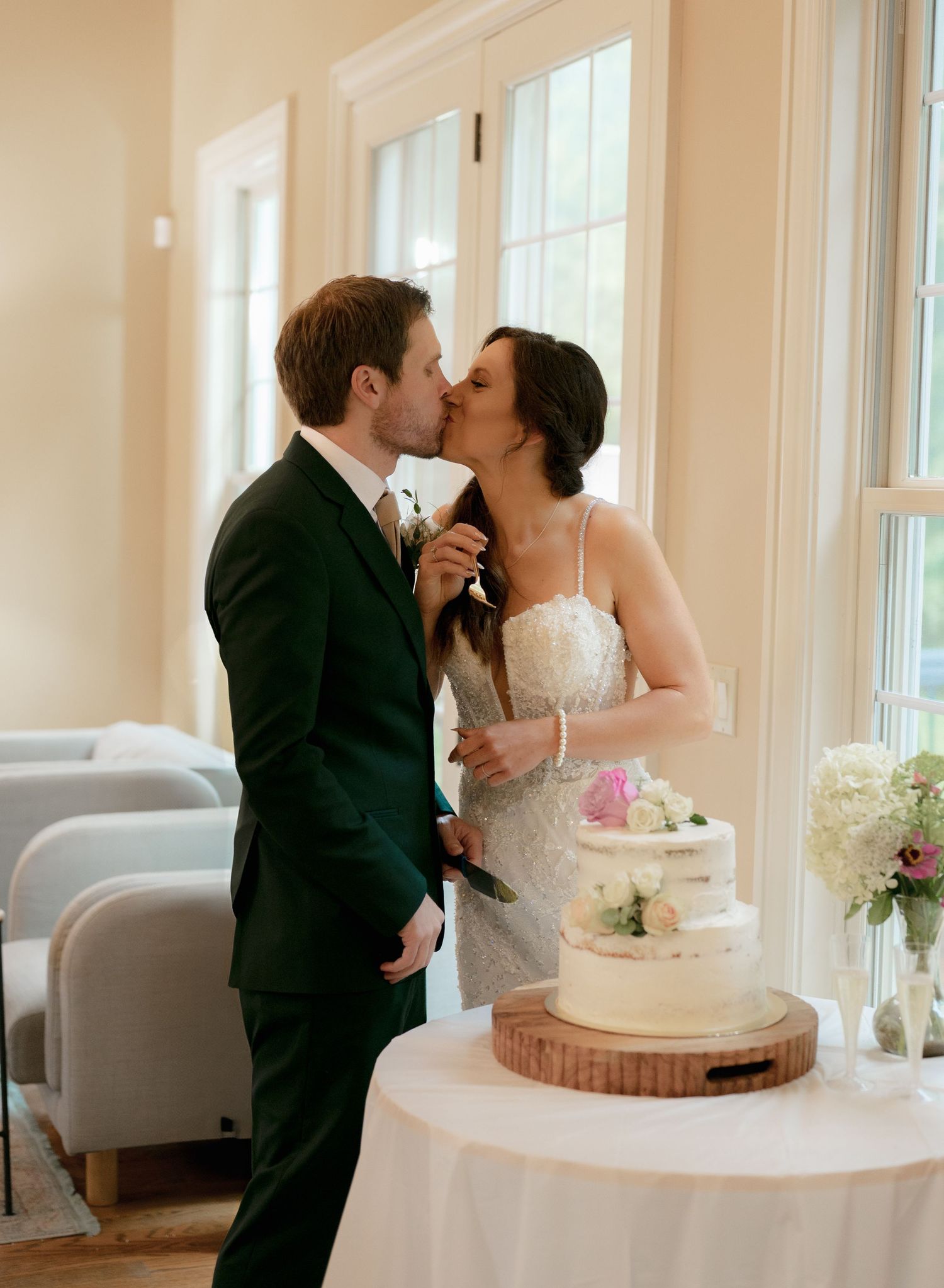 Bride and groom share a sweet kiss beside their elegant semi-naked wedding cake decorated with fresh flowers.