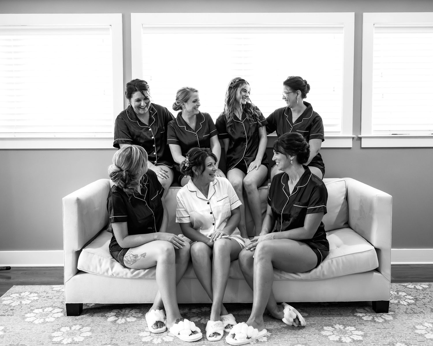 Bridesmaids in black satin pajamas gather around bride in white on a sofa in black and white photo.