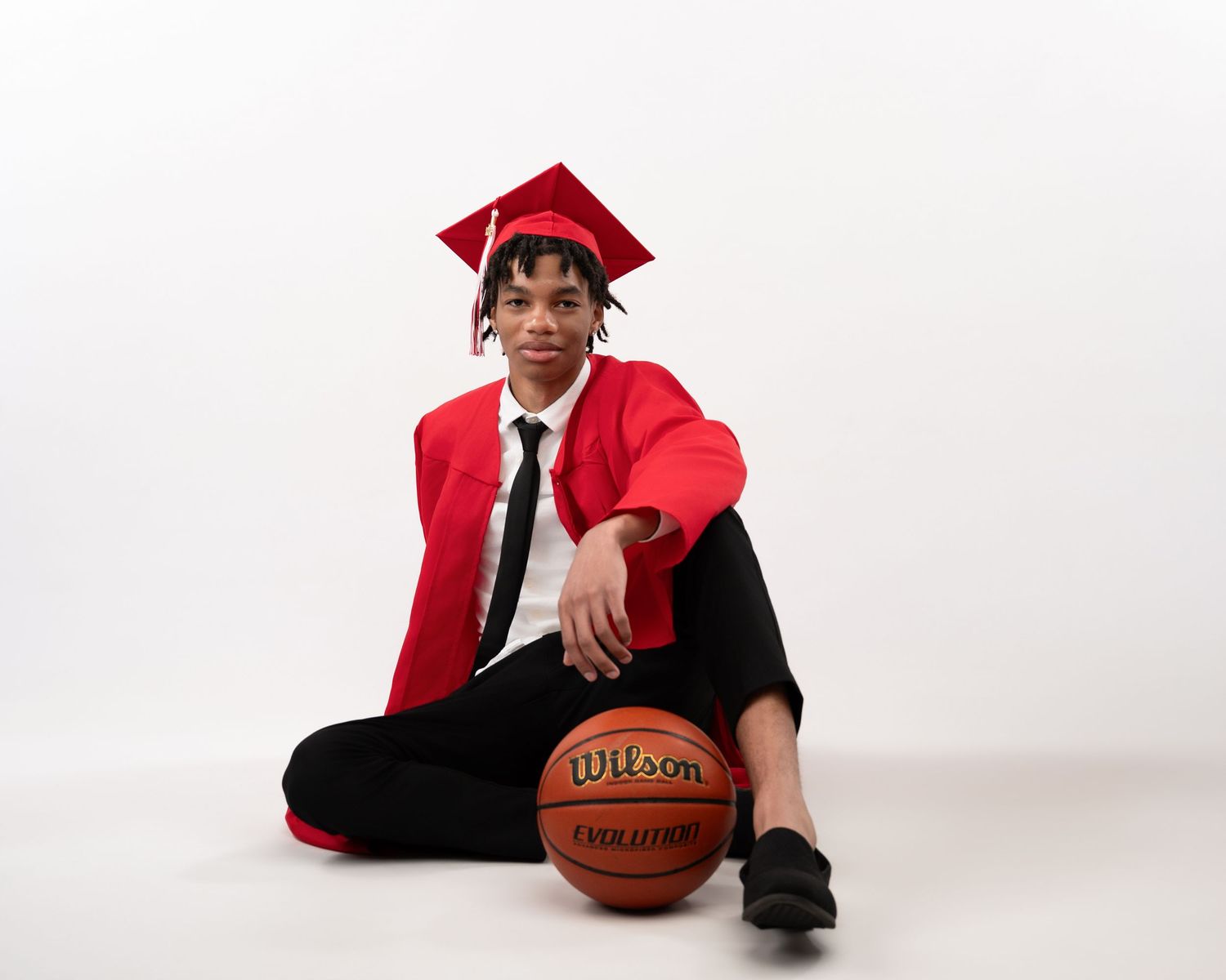 High school graduate in red cap and gown sits casually with Wilson basketball in studio portrait.