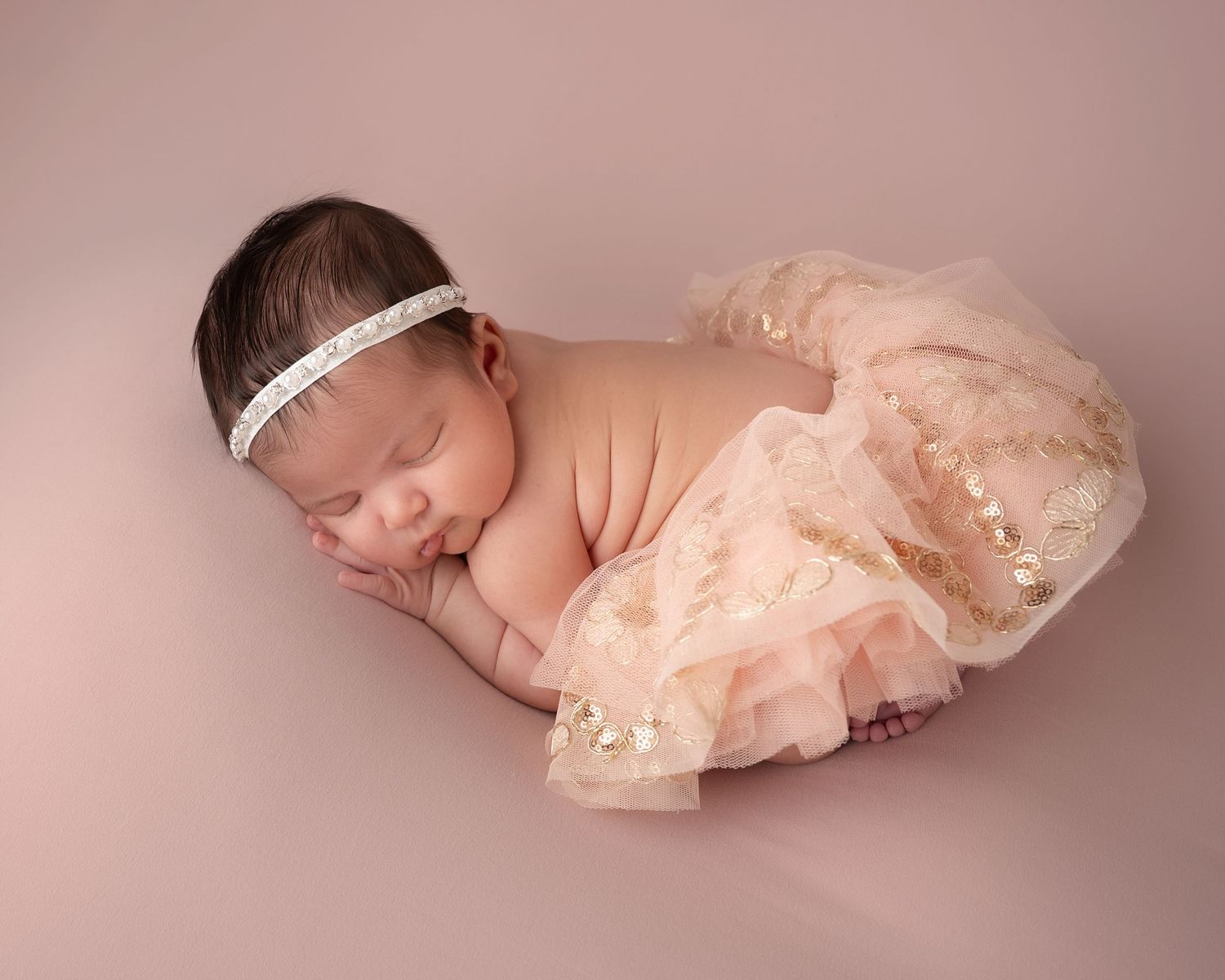 Sleeping newborn baby girl in pink tutu skirt and pearl headband on pink backdrop.