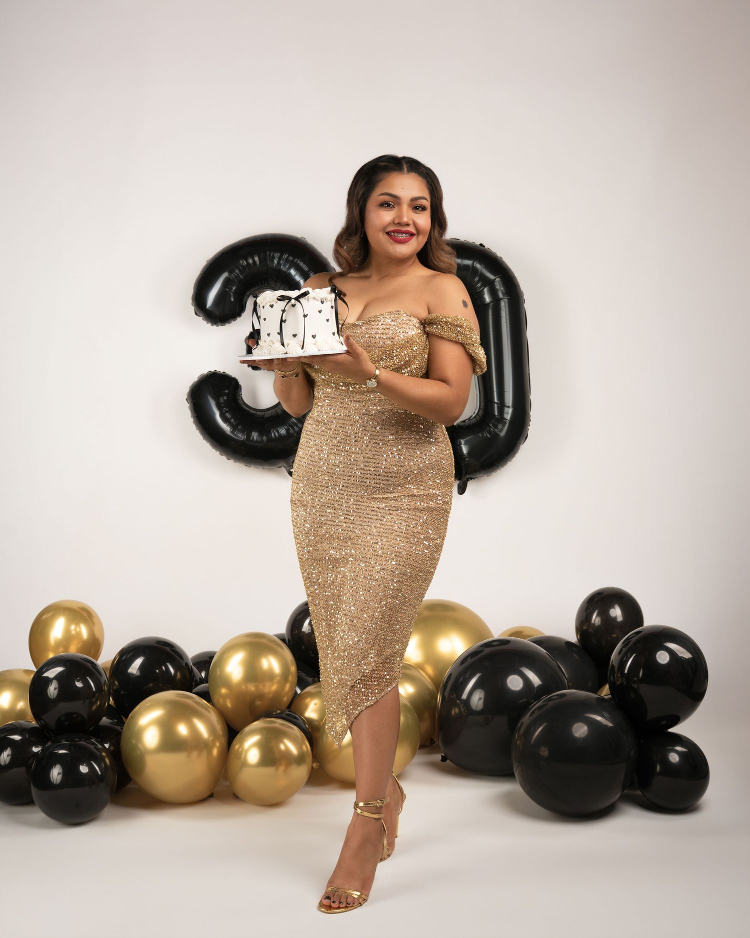 Woman in a sparkling gold off-shoulder dress holds a birthday cake beside black and gold balloons and a large black number 30 balloon.