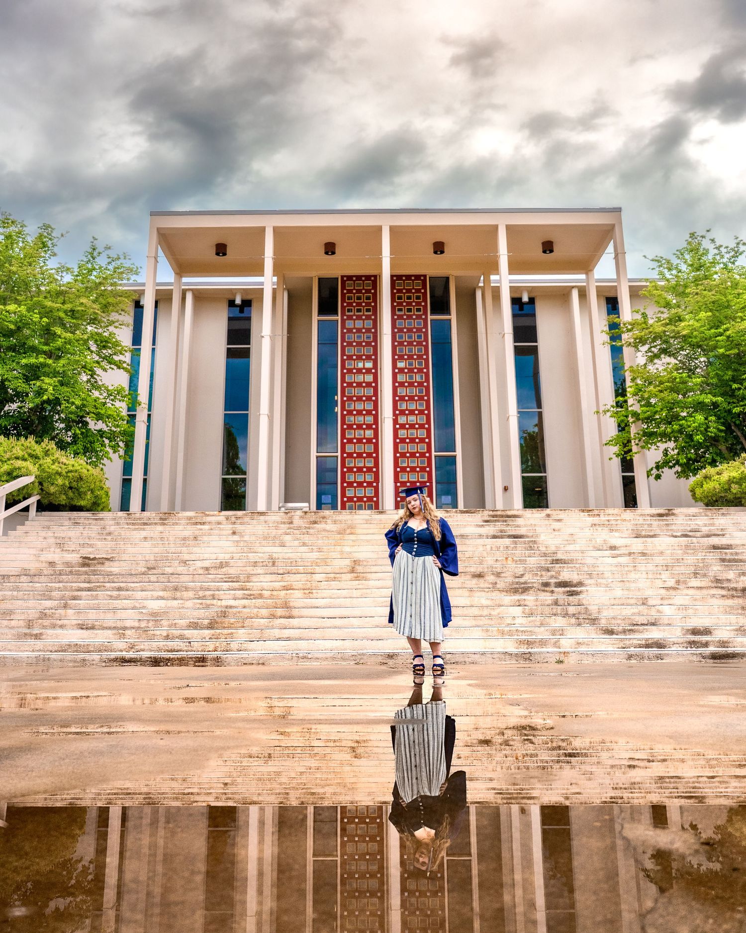 Graduate in blue cap and gown standing before columned building reflected in puddle under dramatic cloudy sky.