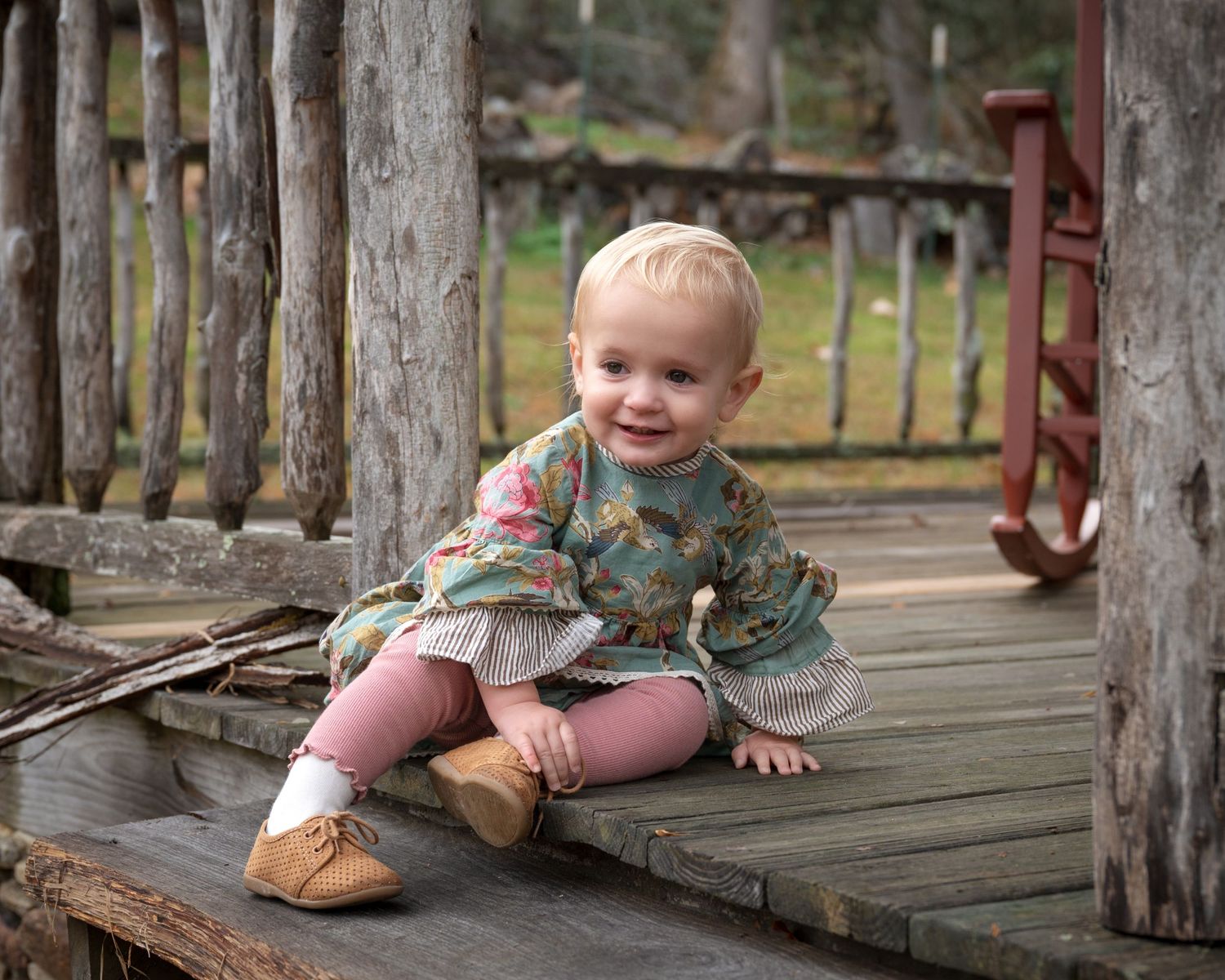 Adorable baby girl in floral dress sitting on wooden bridge deck smiling at camera outdoors.