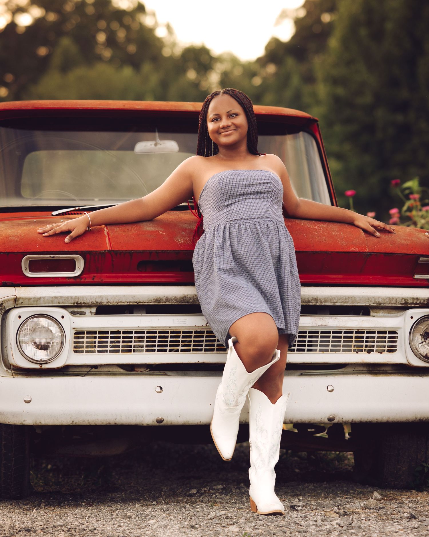 Smiling young woman in denim dress and white cowboy boots leaning against a vintage red truck.