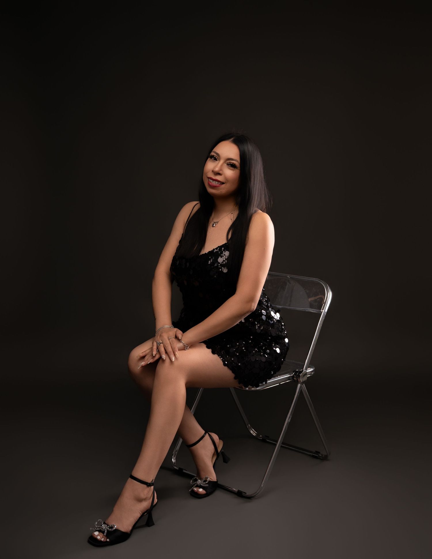 Woman with long dark hair sits on a clear acrylic chair in a black sequin dress and strappy heels against a dark studio backdrop.