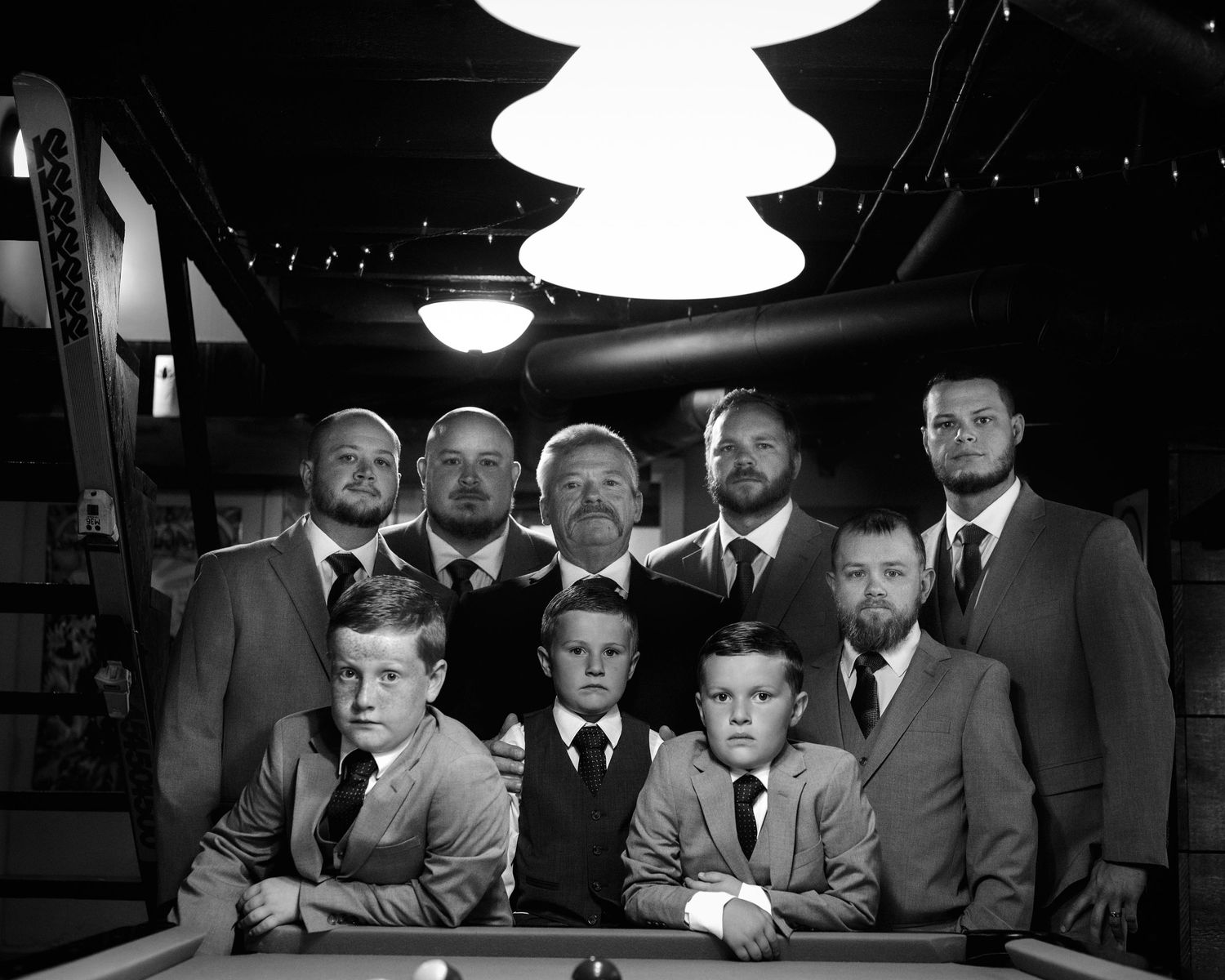 Black and white group portrait of groomsmen and young boys posing around a pool table indoors.
