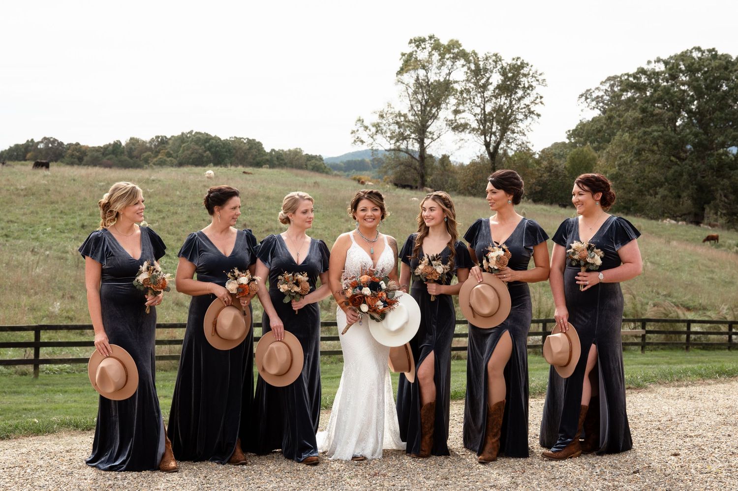 Bridesmaids in navy dresses hold straw hats beside bride in white gown at an outdoor farm wedding.