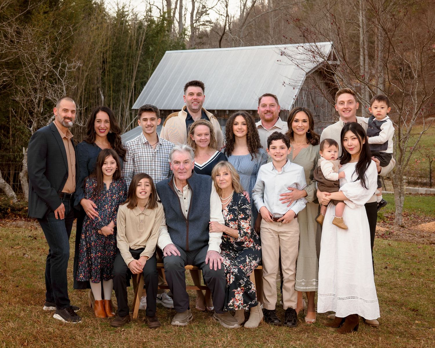 A large extended family poses together outdoors near a rustic barn during a winter family portrait.