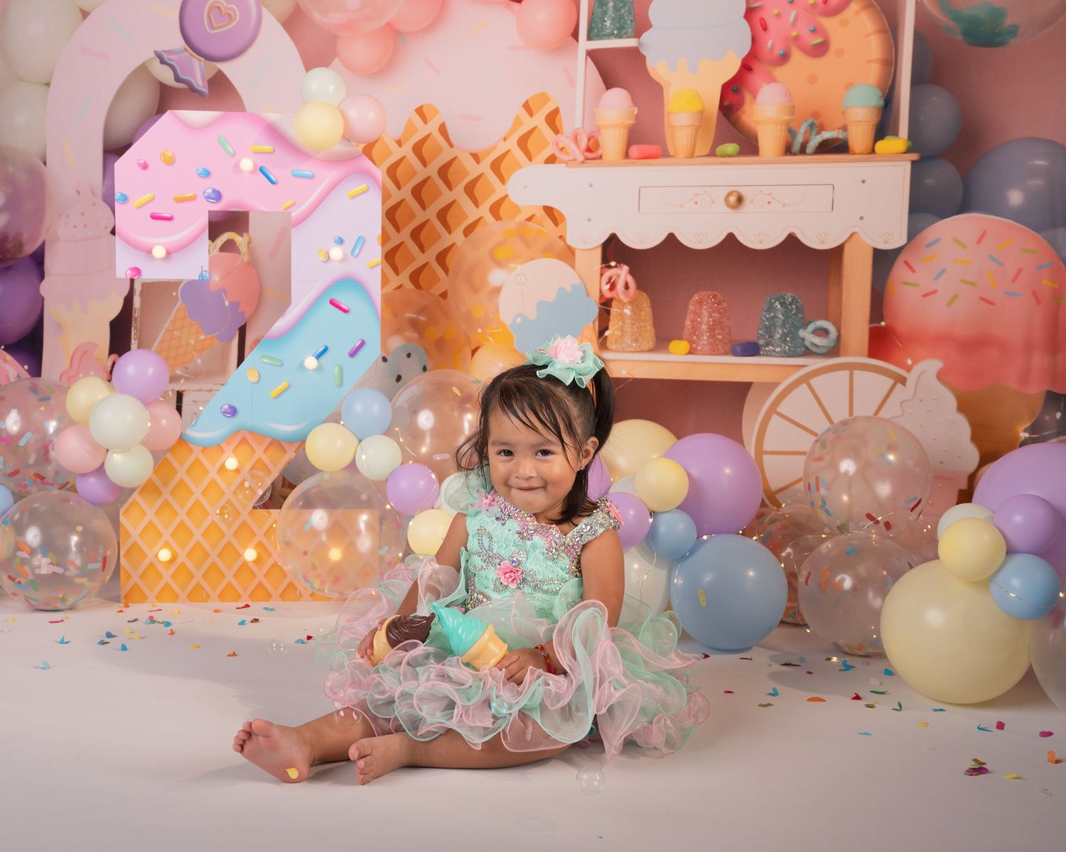 Smiling toddler girl in mint dress celebrates birthday surrounded by pastel ice cream themed balloons and decorations.