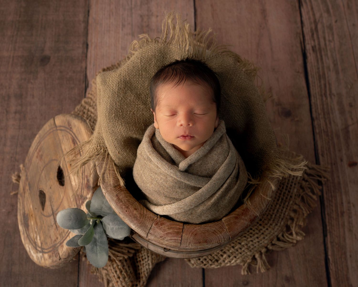 Sleeping newborn wrapped in tan burlap swaddle nestled in rustic wooden spool on dark wood floor.