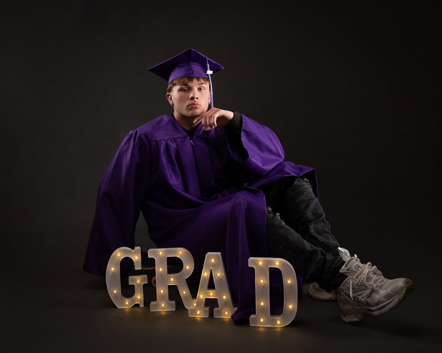 Graduate in purple cap and gown sits beside illuminated GRAD marquee letters in dark studio.