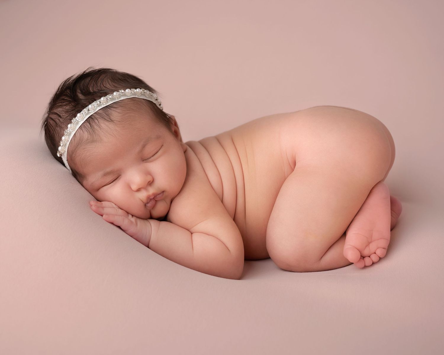 Peaceful sleeping newborn baby girl posed on a pink blanket wearing a delicate rhinestone headband.