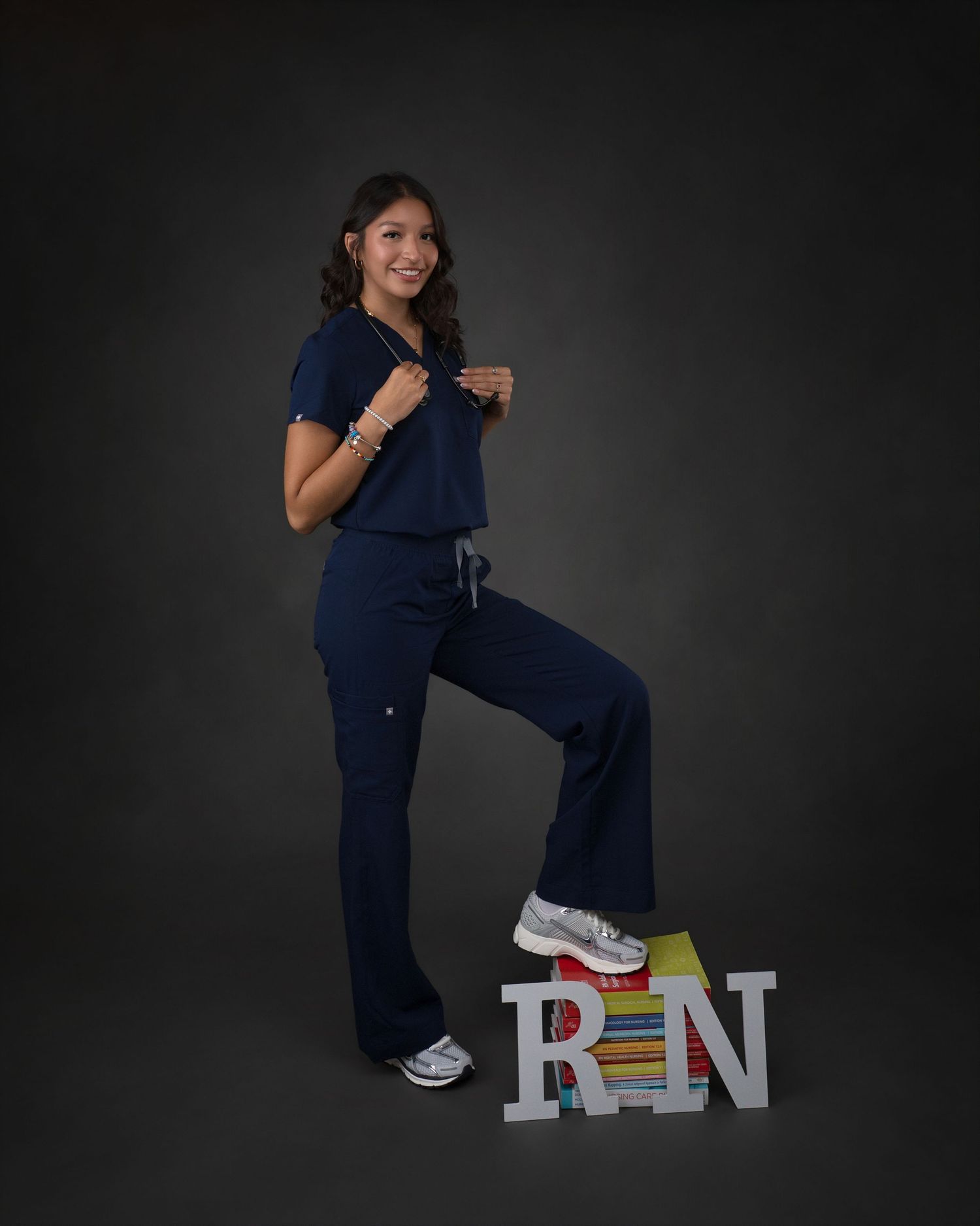 Woman in navy scrubs posing with RN letters and stacked books for nursing graduation portrait.