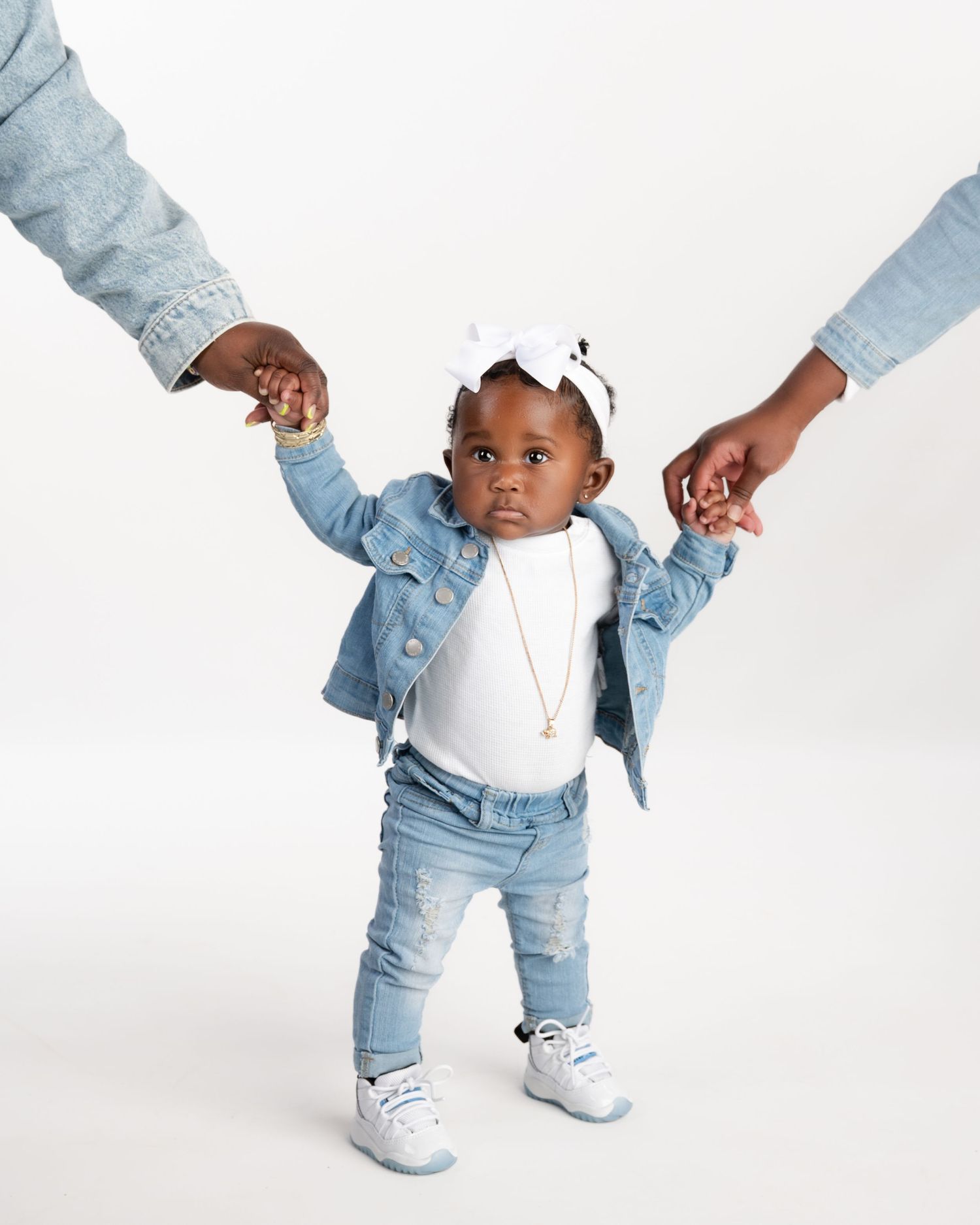 Adorable baby girl in denim outfit stands holding parents hands in bright studio photo.