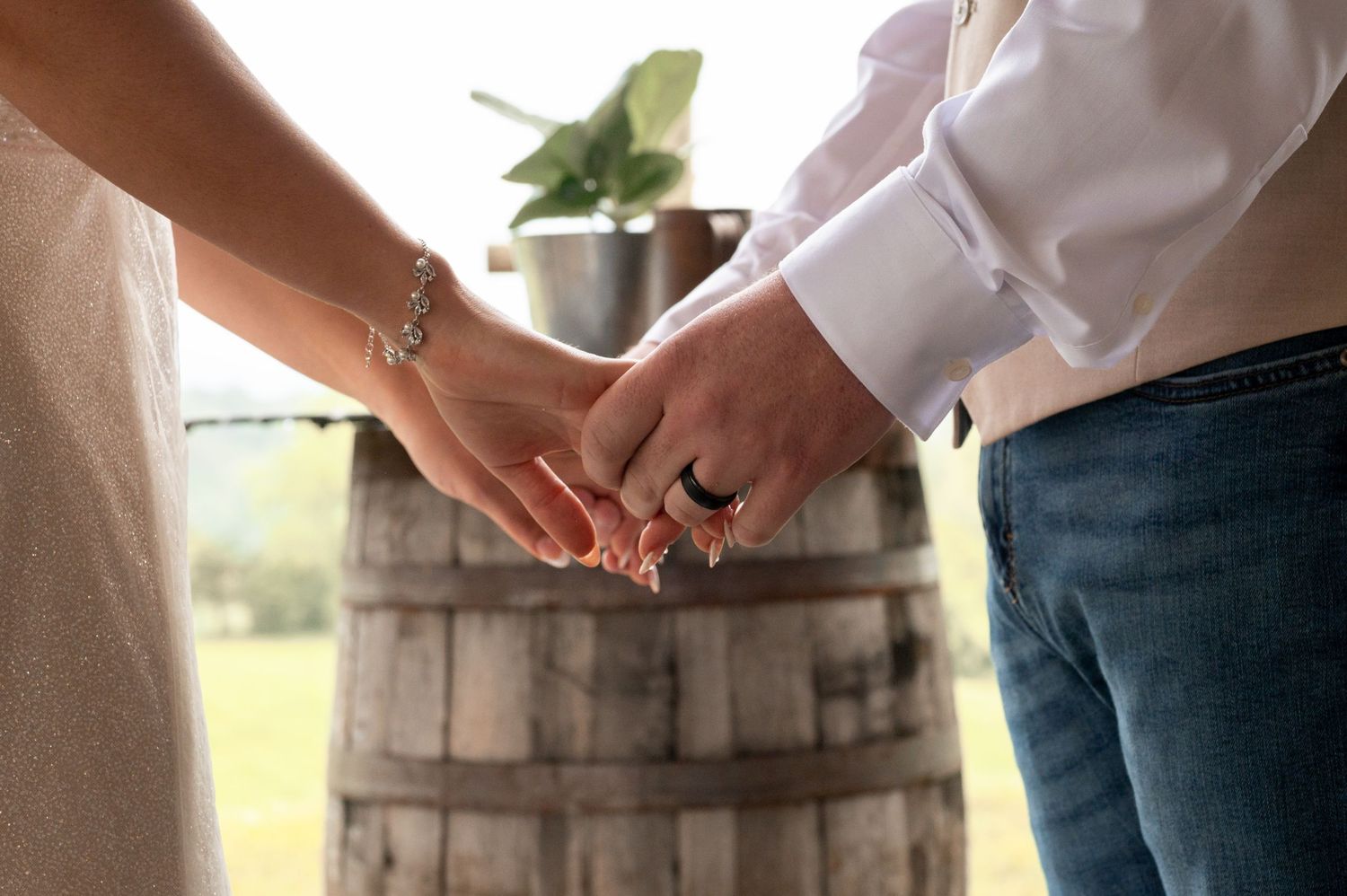 Bride and groom holding hands near a rustic wooden barrel at an outdoor wedding ceremony.