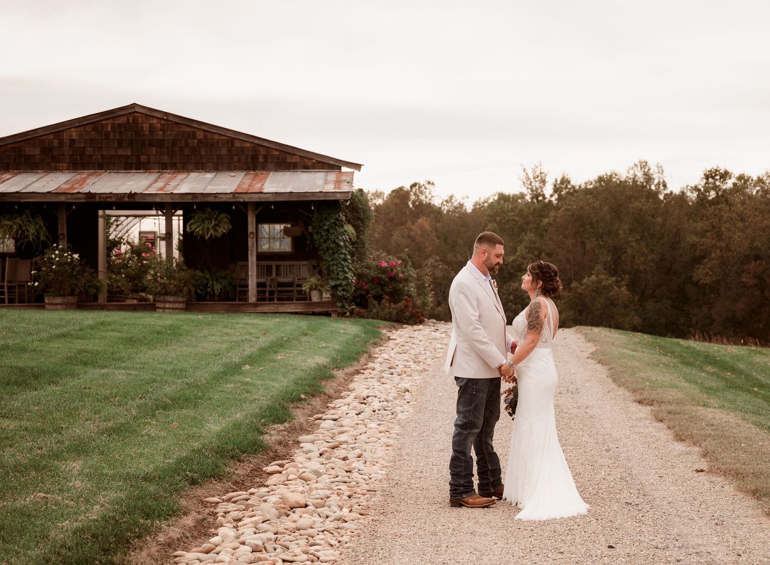Bride and groom hold hands on a gravel path beside a rustic barn at sunset.